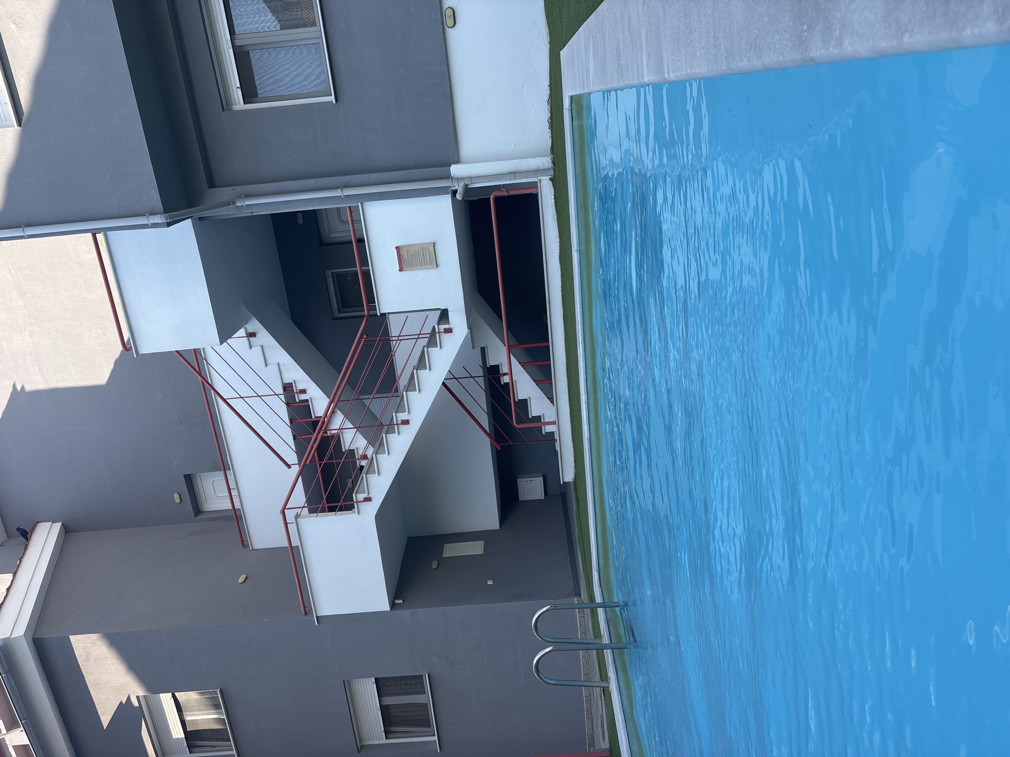 The image depicts a swimming pool in front of a multi-story building with an external staircase. The pool is filled with bright blue water and has a light gray edge. In the foreground, the top part of two silver pool ladders is visible. Behind the pool, there is a patch of artificial green turf. The building is painted in a dark gray color with white accents on the balconies and the external staircase. The staircase has red railings. The building has multiple windows with white roller blinds. The sky is not visible. The overall scene is outdoors, under natural light, suggesting it is daytime. The location is identified as Nea Irakleitsa, Greece.