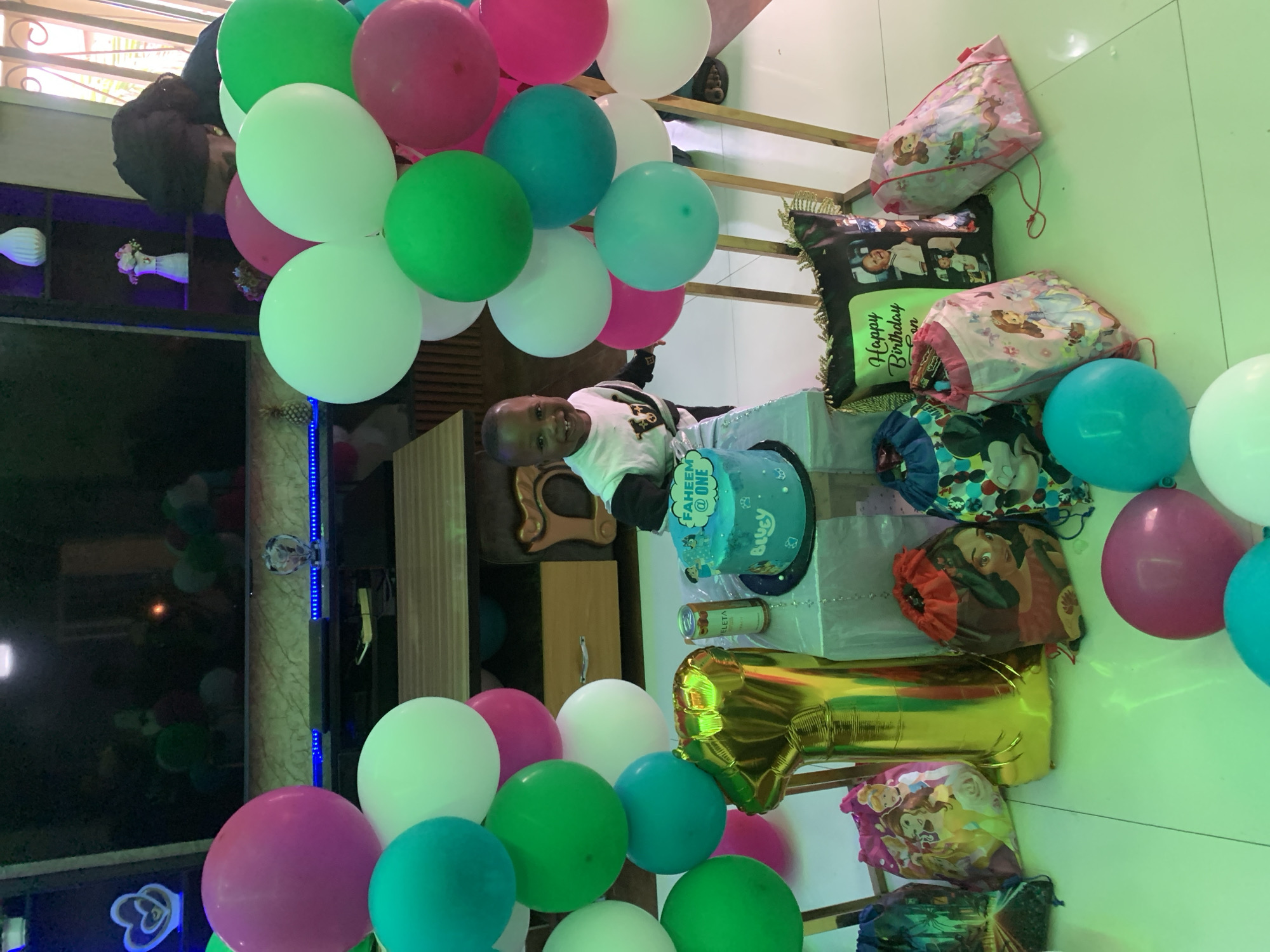 An indoor birthday celebration is captured in the city of Jos, Nigeria. The scene is dominated by colorful balloons and party items. In the center-left, a young Black child, identified as "FAHEEM" by a cake topper, stands behind a small table, smiling broadly and looking directly at the camera. He appears to be a toddler, likely celebrating his first birthday, as indicated by the cake topper "FAHEEM @ ONE" and a large gold foil balloon shaped like the number "1" resting on the floor to his left. He wears a white long-sleeved shirt with a black collar and dark detailing on the chest. His left hand rests on the edge of the table. On the table is a two-tier blue birthday cake with white frosting accents. The cake topper reads "FAHEEM @ ONE," and the side of the cake has the name "Bluey" inscribed. Beside the cake, a silver "CELETA" beverage can is visible. The table is covered with a white cloth. The room is extensively decorated with clusters of balloons in teal, green, white, and pink, arranged on stands. One cluster is positioned to the left of the cake table, and another larger cluster is to the right. Additional loose balloons in pink, teal, and white are scattered on the light-colored, reflective floor in the foreground. To the far right, partially obscured by a balloon cluster, an adult Black individual is visible from the top of the head and shoulder. They have dark, braided hair and are wearing a dark top. Their action is not fully discernible, but they appear to be leaning forward. Scattered on the floor in the foreground are several drawstring bags featuring various cartoon characters, including Disney Princesses (Ariel, Belle, Cinderella), Moana, Mickey Mouse, and Princess Sofia. Two decorative cushions are also on the floor to the right: one black with gold fringe, inscribed "Happy Birthday Son" and displaying a photo collage of a baby; the other black with an image of a cartoon character. The background features home furnishings. A large flat-screen television is mounted above a dark entertainment unit, which has blue LED strip lighting running along its length. Reflections of balloons and room lighting are visible on the TV screen. Various decorative items are on the entertainment unit, including a glowing heart ornament, a small clear glass ornament, and a miniature pineapple sculpture. To the right of the TV setup, a dark