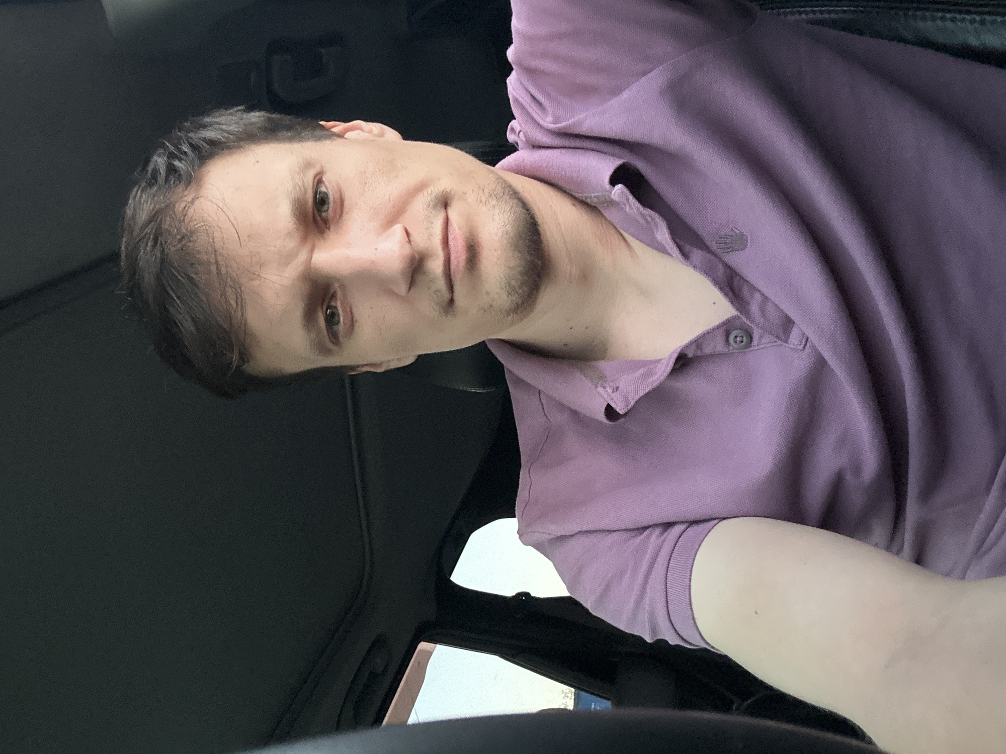 A medium shot selfie captures a man from the chest up, seated inside a vehicle. He has light skin, short brown hair, and a slight stubble. He is wearing a purple polo shirt with a collar and two buttons at the placket. A small, dark logo, possibly a handprint, is visible on the left side of his chest. The background is dark, suggesting the interior of the car, with a hint of the sky and window on the left. The man is looking directly at the camera with a neutral expression. The image appears to be taken during daylight. There is no information in the image to indicate the location in Shatava, Ukraine.