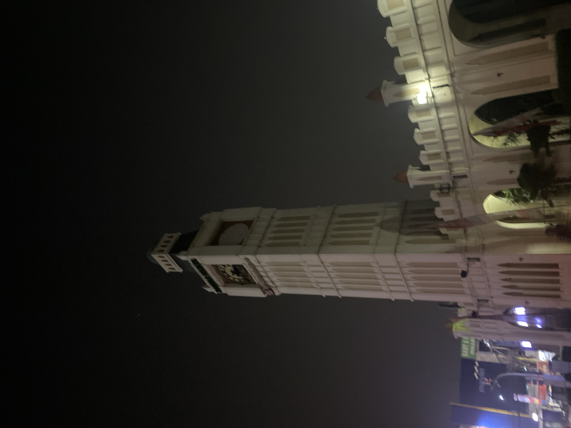 An outdoor, low-light, wide-angle shot captures a replica of the Elizabeth Tower, commonly known as Big Ben, at night in the city of Tangerang, Indonesia. The tower, a prominent white structure with a dark roof, stands against a dark, hazy sky. Its clock face is visible, though not illuminated. To the right of the tower is a white, crenellated wall with arched openings, suggesting a larger complex. Flags, one appearing red and white, are displayed on the wall. In the background, traffic lights and signs, including one that reads "BABY'S PHARMA," are dimly visible, indicating an urban setting. A few street lamps cast a faint glow on the scene. The image is slightly blurry, characteristic of night photography with minimal lighting.