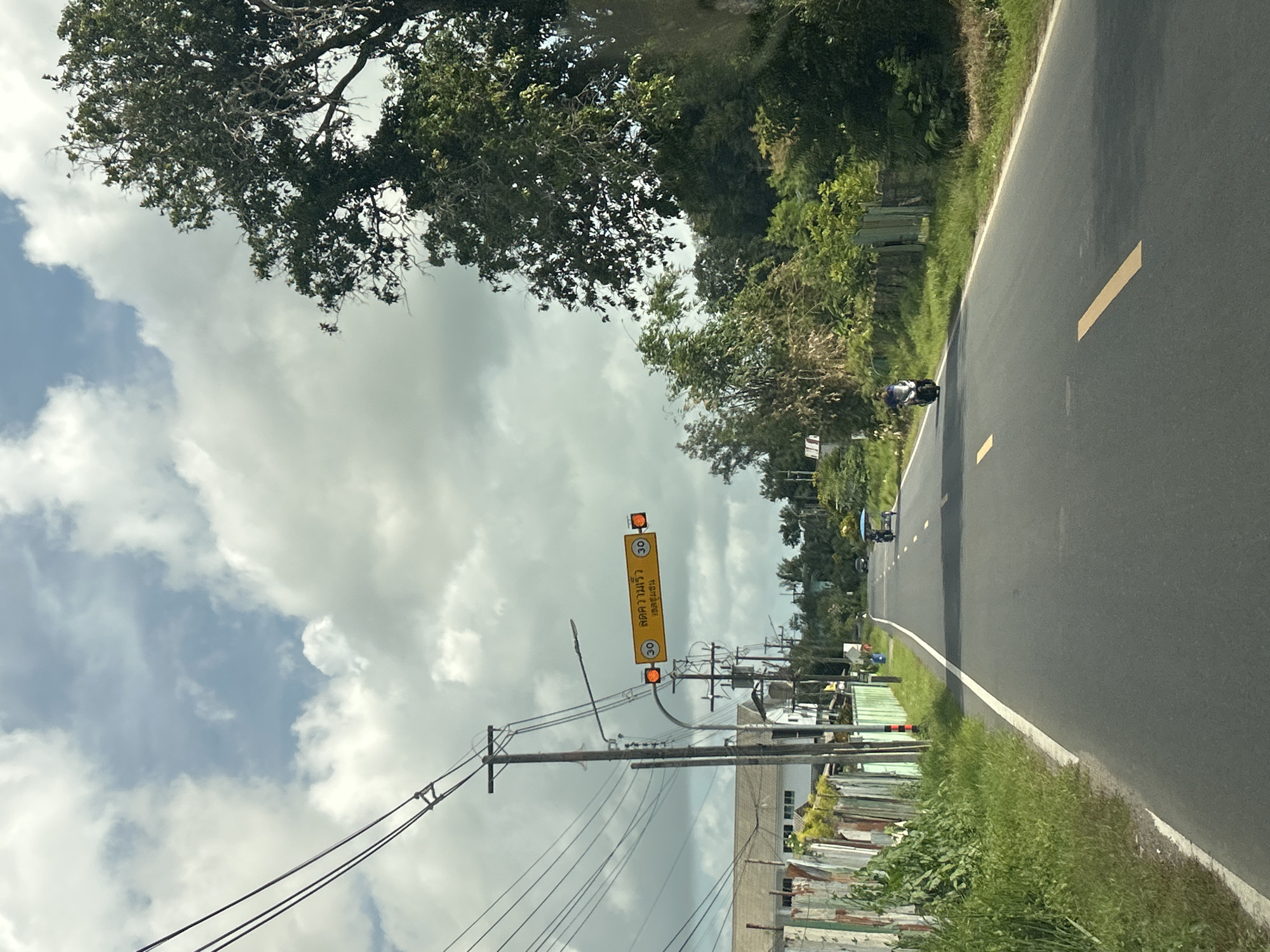 In Ratsada, Thailand, a two-lane asphalt road is depicted under a partly cloudy sky. The road features yellow painted lines as lane dividers. On the left side of the road, a weathered metal fence runs along a grassy embankment, with utility poles and power lines overhead. Beyond the fence, buildings and vegetation are visible. On the right side of the road, a line of trees borders the pavement. In the distance, two motorcycles are traveling on the road; one is a scooter with a blue canopy, and another is a motorcycle carrying a single rider. A speed limit sign with the number "30" in a yellow circle is displayed on an overhead structure, accompanied by flashing orange lights, indicating a speed limit of 30 kilometers per hour.