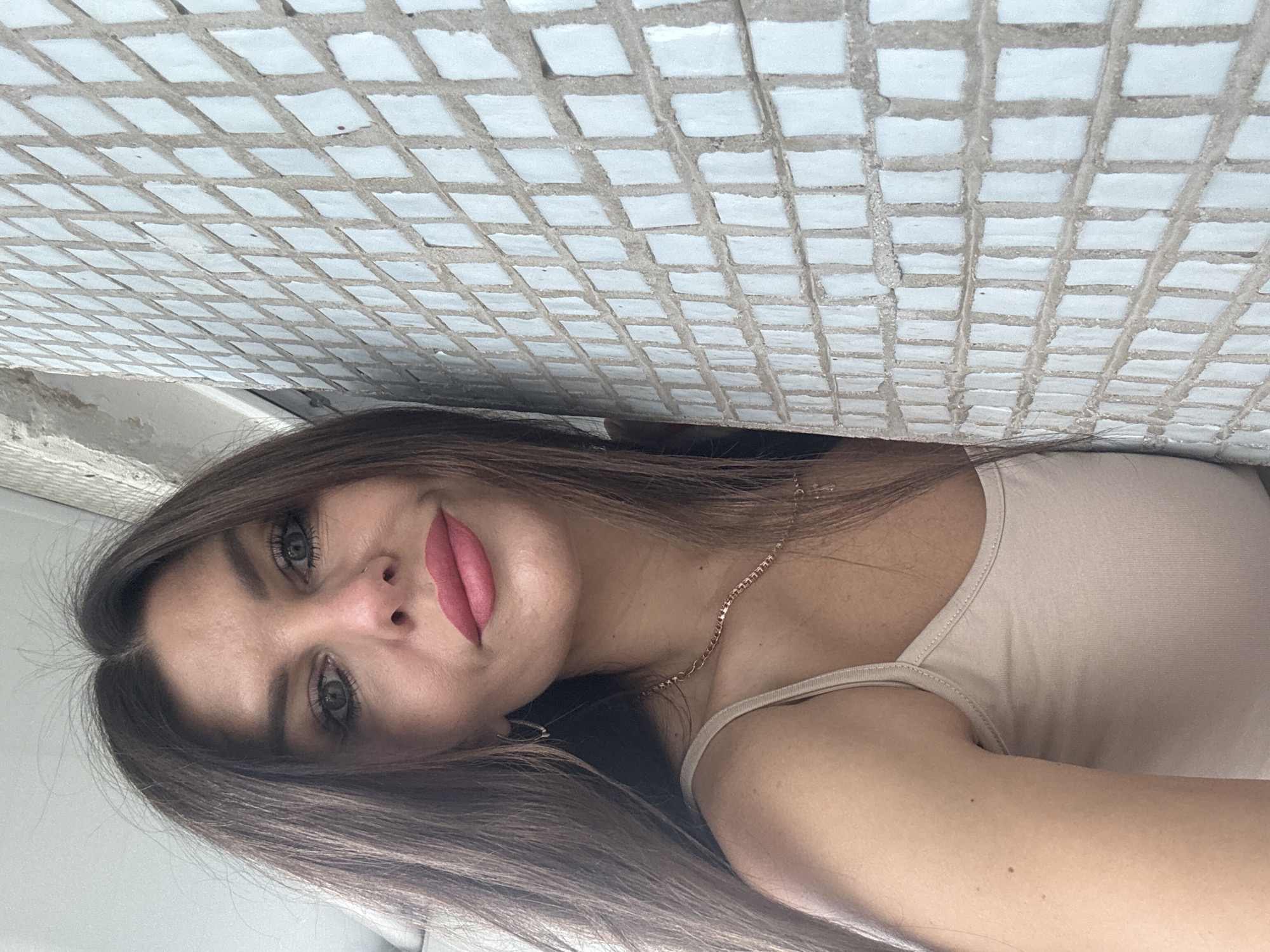 A woman with long, dark brown hair is leaning against a wall with a mosaic of small, light blue tiles. She is wearing a light beige spaghetti strap top and a delicate gold chain necklace. Her makeup includes pink lipstick, eyeliner, and mascara. She has a neutral expression and is looking directly at the camera. The environment appears to be outdoors, possibly on a balcony or a side of a building, with rough concrete structures visible in the background. The lighting suggests it is daytime. There is no discernible information about the specific location within Chaikovsky, Russia, beyond the fact that it is likely an urban or semi-urban setting.