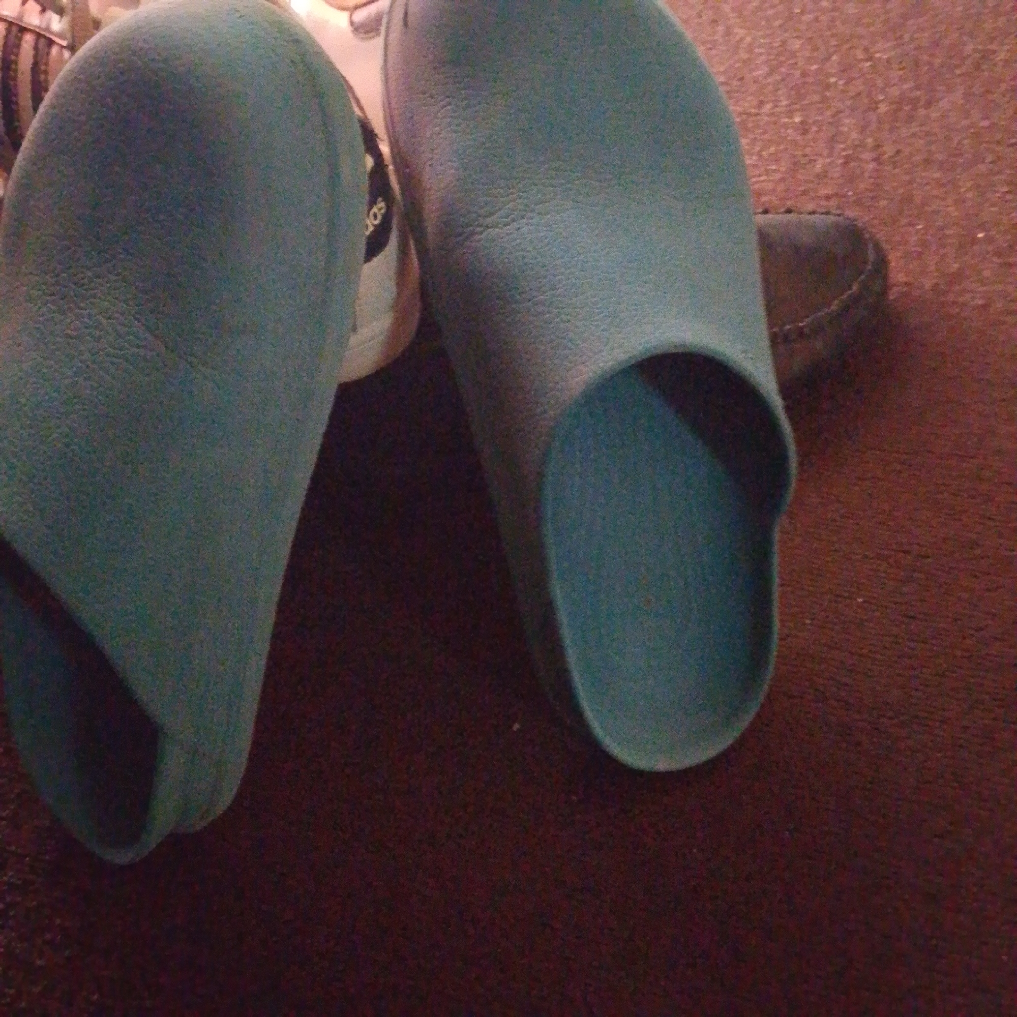 The image shows two teal-colored slip-on shoes placed on a dark brown carpeted surface. The shoes appear to be made of a soft material and have a rounded toe. One shoe is positioned on the left side of the frame, facing slightly towards the right. The other shoe is on the right side, also facing towards the left. In the background, partially visible, is a portion of another shoe or object with the word "JOS" printed on it in black lettering. The lighting in the image is dim, casting shadows and creating a slightly grainy texture. The scene is indoors, likely a residential setting. There are no people visible in the image. The context of Jalingo, Nigeria, is not evident from the visual elements of the photograph.