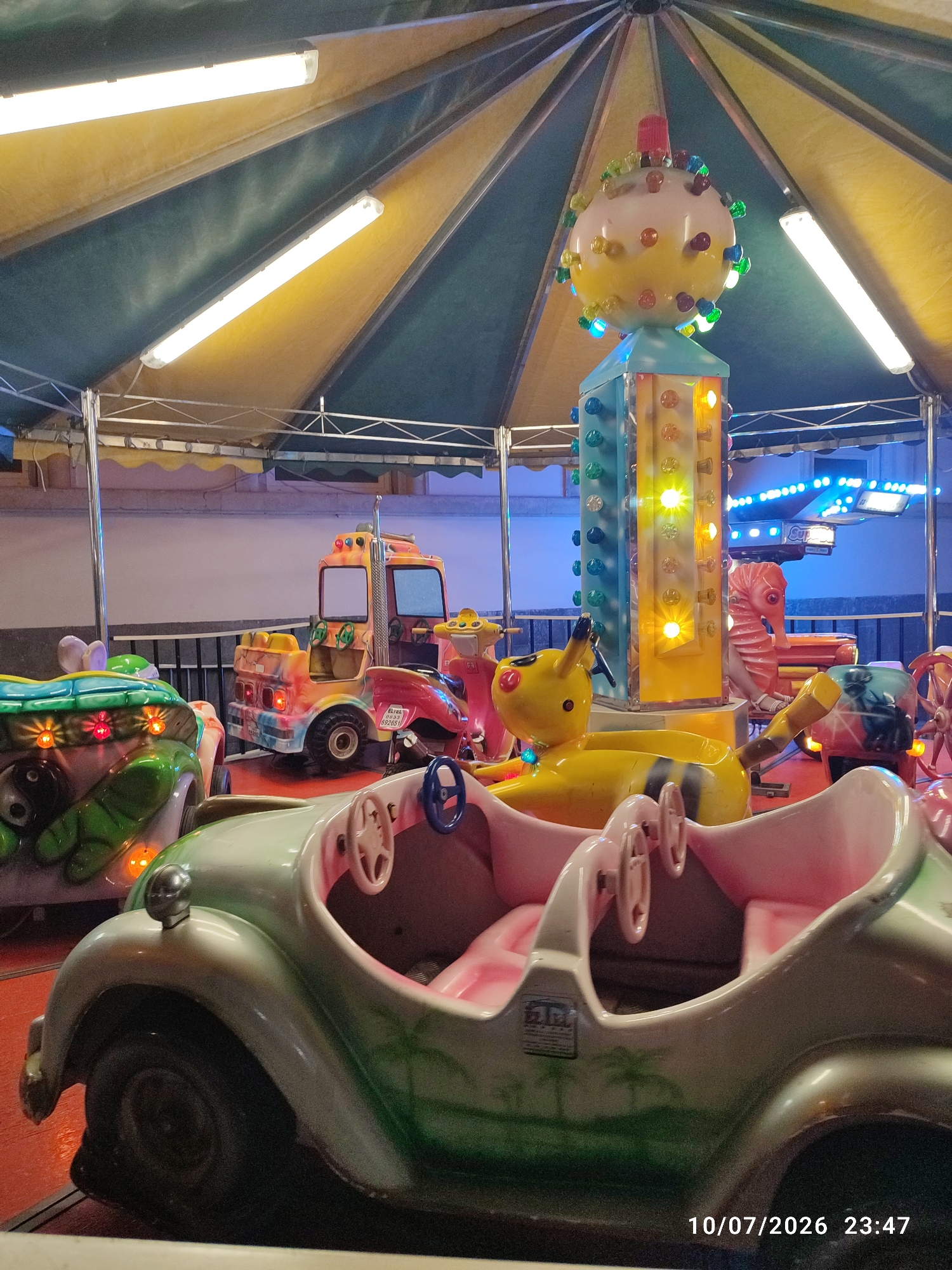 The image shows a carousel in Militello in Val di Catania, Italy. The carousel features several ride-on vehicles, including a light green retro-style car in the foreground with pink seating and palm tree decals. Behind it are other colorful attractions such as a yellow bumblebee-shaped ride, a red and orange truck, and a seahorse-themed ride. The carousel is covered by a tent with alternating yellow and dark green sections, illuminated by fluorescent lights. The date stamp on the bottom right corner indicates the photo was taken on October 7, 2026, at 23:47. There are no people visible in the image.