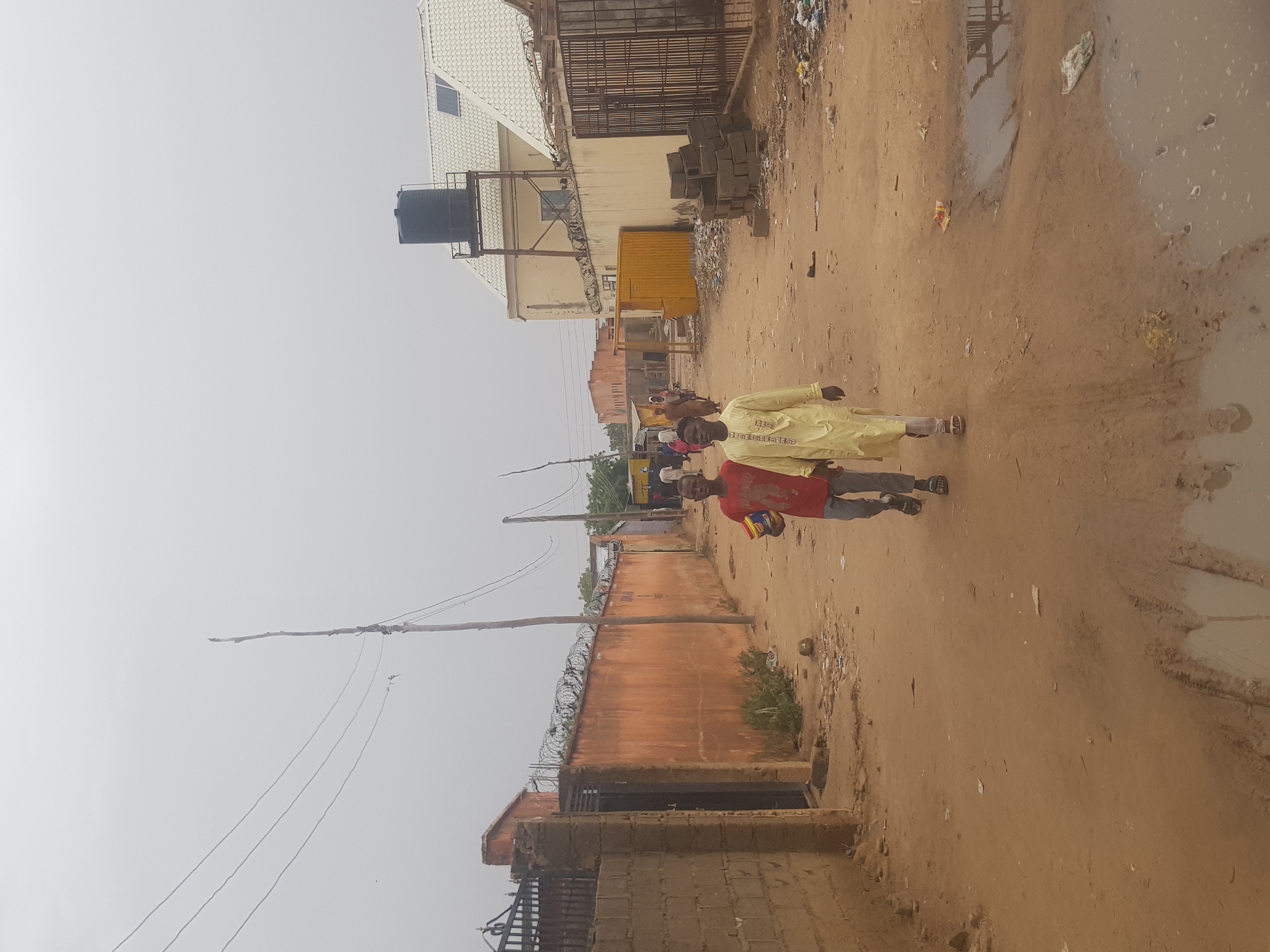 The image shows a dirt road in Dabawa, Nigeria. The road is muddy in places. Two people, one wearing a light yellow robe and the other a red shirt, are walking down the road. A building with a water tank on top is visible in the background. A fence topped with barbed wire runs along one side of the road. Overhead power lines stretch across the sky. There is garbage strewn along the road and near the buildings.