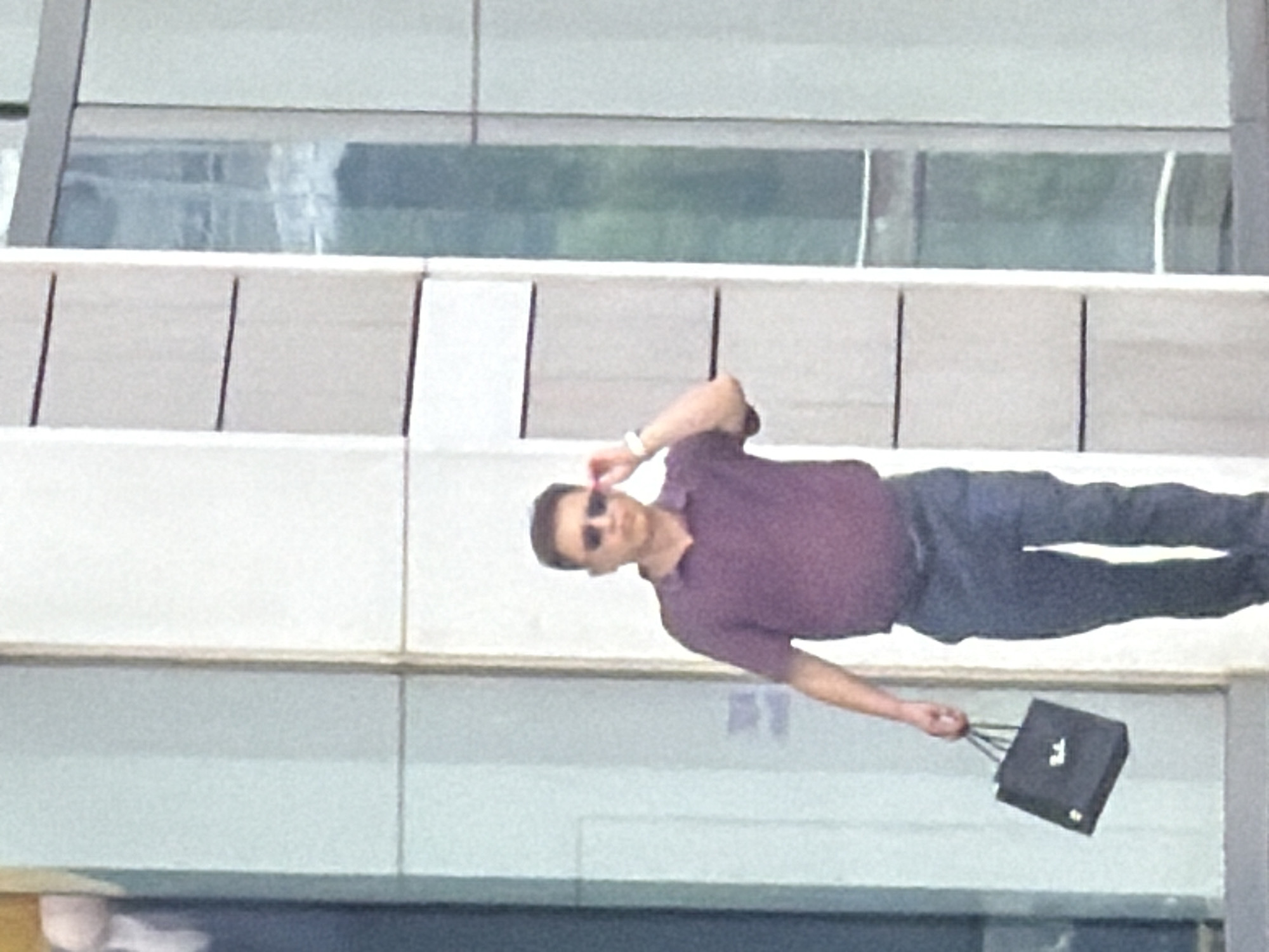 An East Asian man wearing sunglasses and a purple polo shirt walks outdoors while holding a black shopping bag in his left hand. He is walking to the right of the frame. His right hand is raised to adjust his sunglasses. He is walking in front of a building with large windows. The scene is likely in Yuzhong District, China, given the context provided.