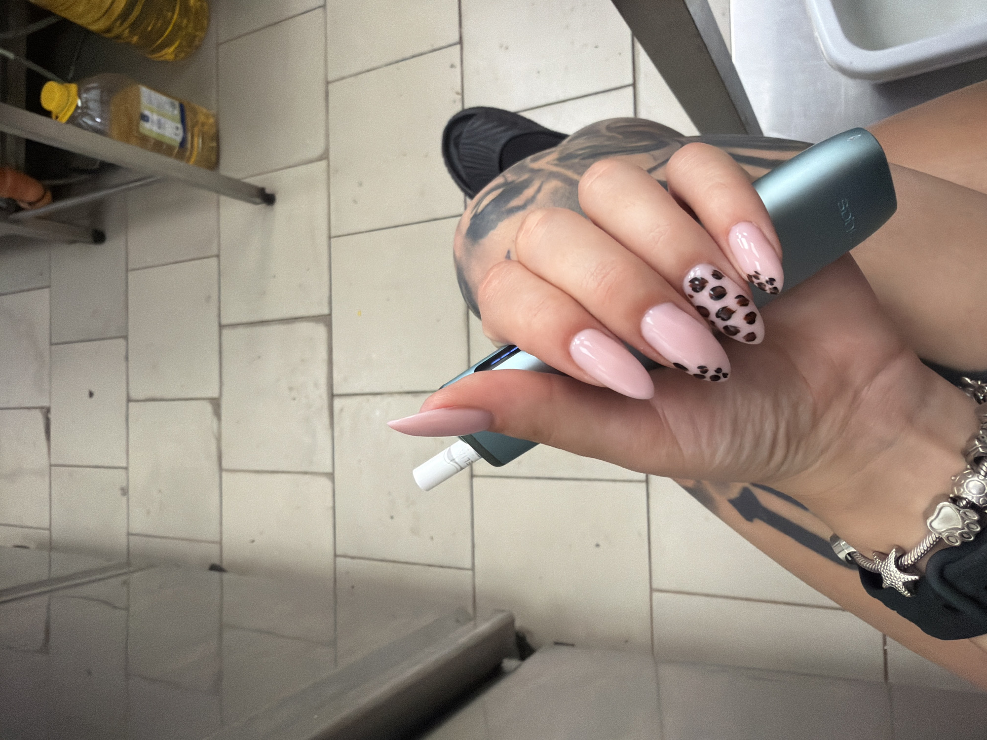 The image displays a close-up view of a person's left hand, adorned with light pink almond-shaped fingernails, two of which feature a leopard print design. The individual is holding a teal-colored electronic smoking device in their hand. Tattoos are visible on the person's arm and hand. A silver charm bracelet with several charms, including a starfish and a paw print, is worn on the wrist.
The background consists of light-colored tiled flooring, characteristic of an indoor environment. Partially visible in the background are legs of a metal structure, possibly shelving or furniture, and two plastic bottles containing yellow liquid, likely cooking oil, suggesting a commercial or storage area. The scene is devoid of specific landmarks identifying it as Plovdiv, Bulgaria. The lighting suggests an indoor, possibly commercial setting, with overhead lighting contributing to the overall illumination. The camera angle is overhead, focusing on the hand and the device.