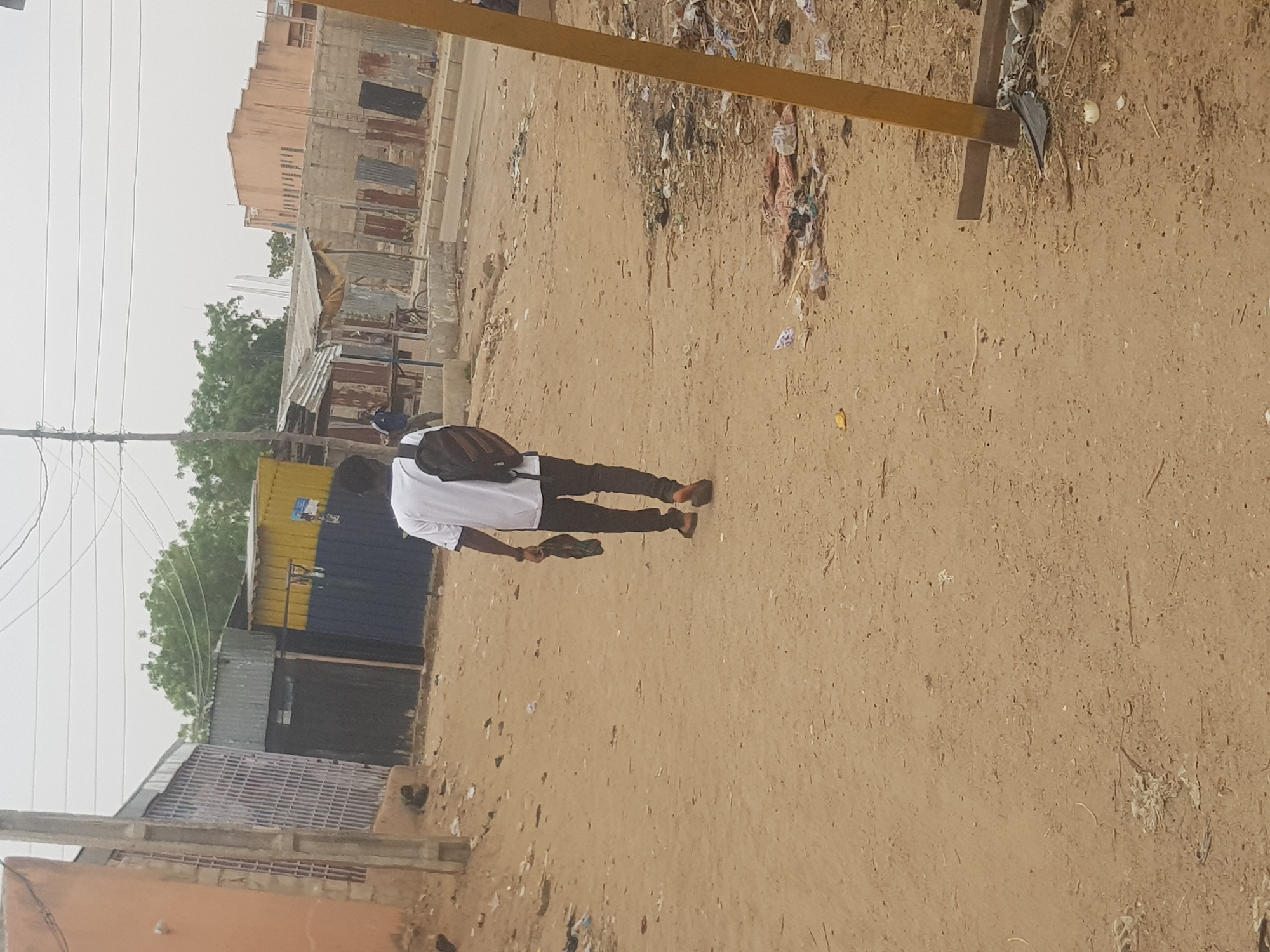 A man in a white shirt, brown pants, and brown shoes is walking on a dirt road. He is carrying a black object in his right hand. He has a backpack on. The road is littered with trash. There is a building in the background with corrugated metal walls. There is a wooden structure in the foreground on the right. There are trees and power lines visible in the distance. The location appears to be a city street in Dutsinma B, Nigeria.
