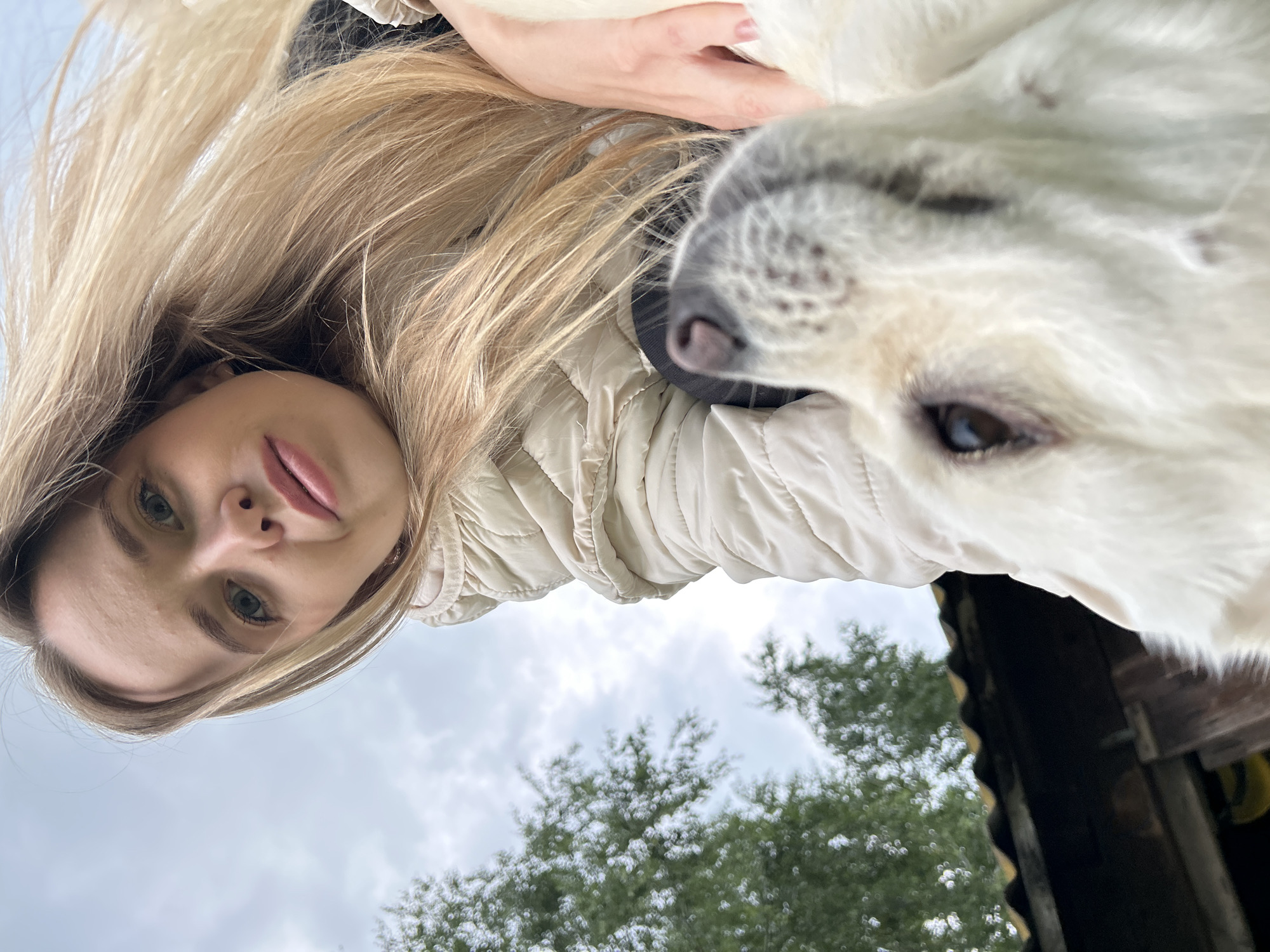 A young blonde woman is taking a selfie outdoors with a white dog in the foreground. The woman is wearing a cream-colored puffer jacket and her hair is falling around her shoulders. The dog, which appears to be a Pomeranian or similar breed, is looking to the left of the frame, with its face partially out of focus. Behind them, a cloudy sky and green foliage are visible, along with part of a dark-colored structure, possibly a roof. The location is not specified in the image.