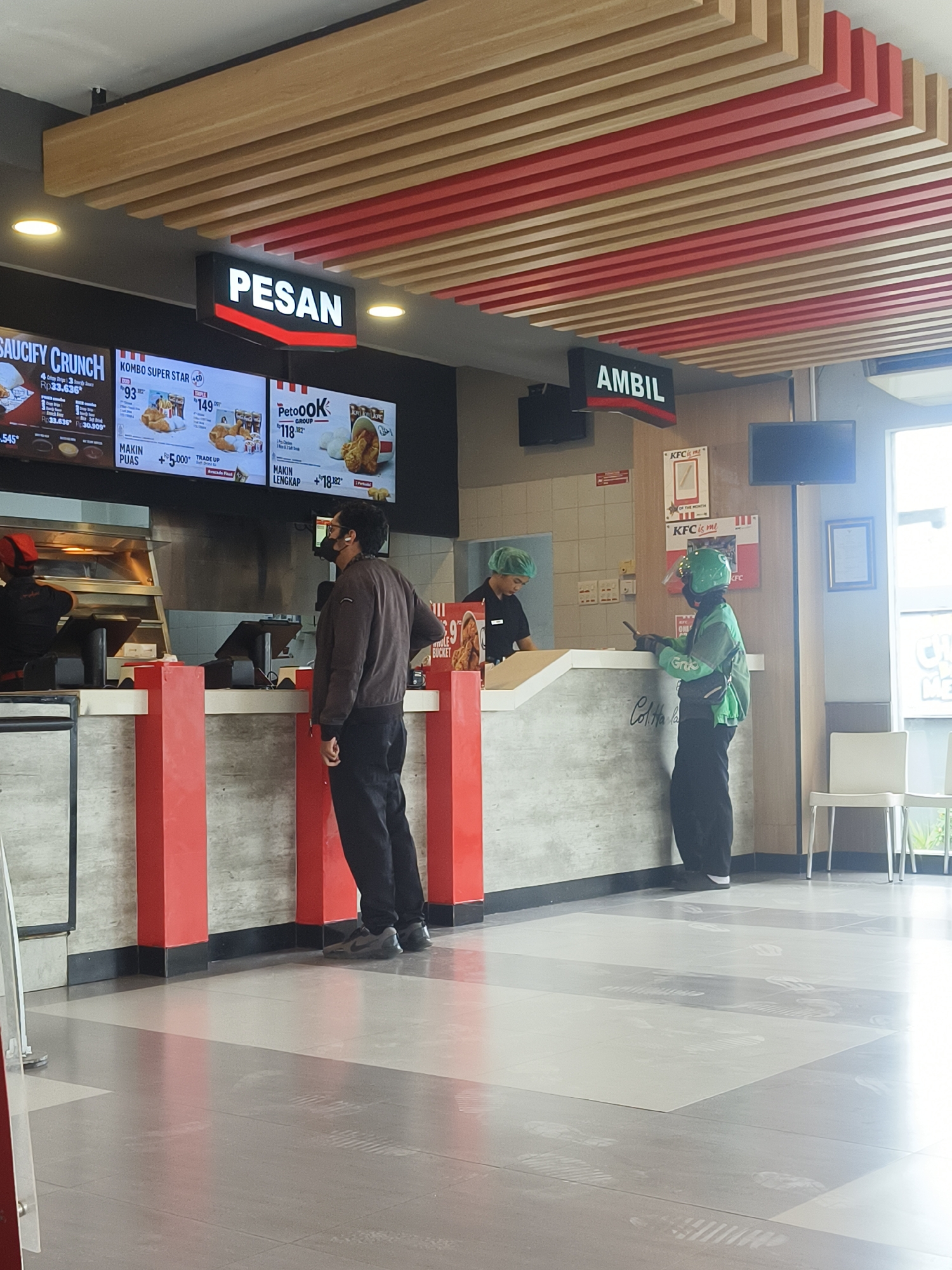 The image depicts the interior of a KFC restaurant, likely located in Denpasar, Indonesia, based on the visual cues of the signage and typical fast-food establishment setup. The scene captures a moment within the restaurant's service area, focusing on the ordering and pickup counters.
In the foreground, a man wearing a brown jacket and black pants stands with his back partially to the camera, observing the counter. He has a black face mask on and appears to be waiting. To his left, two KFC employees are visible behind the counter. One, wearing a red cap and dark shirt, is preparing food. Another employee, wearing a green hairnet and dark shirt, is also behind the counter, seemingly interacting with a customer.
In the middle ground, a delivery driver, identifiable by their green jacket with a logo and a helmet with a visor, stands at the counter holding a smartphone. They appear to be picking up an order. The counter area is marked by red and grey pillars, and the floor is tiled with a pattern of light and dark grey squares, showing visible footprints.
Above the counter, illuminated signs display menu items and instructions. A sign with "PESAN" (Order) is prominently displayed, and another sign with "AMBIL" (Pick Up) is visible further down. Digital menu boards showcase various KFC meal deals, including "SAUCIFY CRUNCH" and "KOMBO SUPER STAR."
The restaurant's ceiling features a distinctive design of slatted wood and red accents, creating a modern aesthetic. In the background, more seating areas and KFC branding are visible, including posters with the slogan "KFC is me." The overall environment is brightly lit, suggesting it is daytime or the interior lighting is strong. The scene captures a typical operational flow within a fast-food restaurant, with customers ordering, staff serving, and delivery personnel collecting orders.