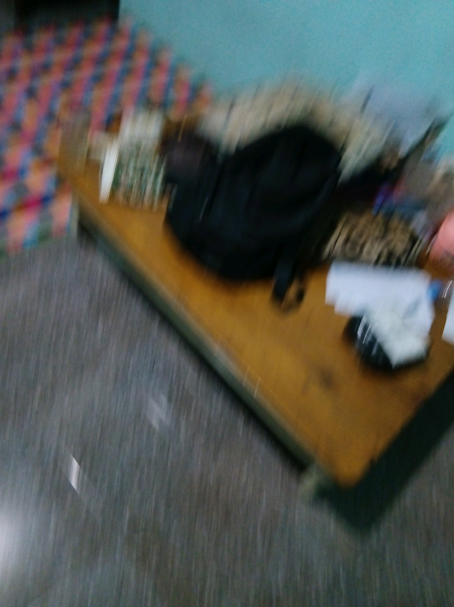 The image depicts a cluttered table or surface with a checkered mat on the left and a blue wall in the background. On the table, there is a black backpack, a white piece of paper, and what appears to be a plastic bag filled with a white substance. The floor is a speckled gray. The image is blurry and does not contain enough detail to identify any specific location in Suka Raya, Indonesia.