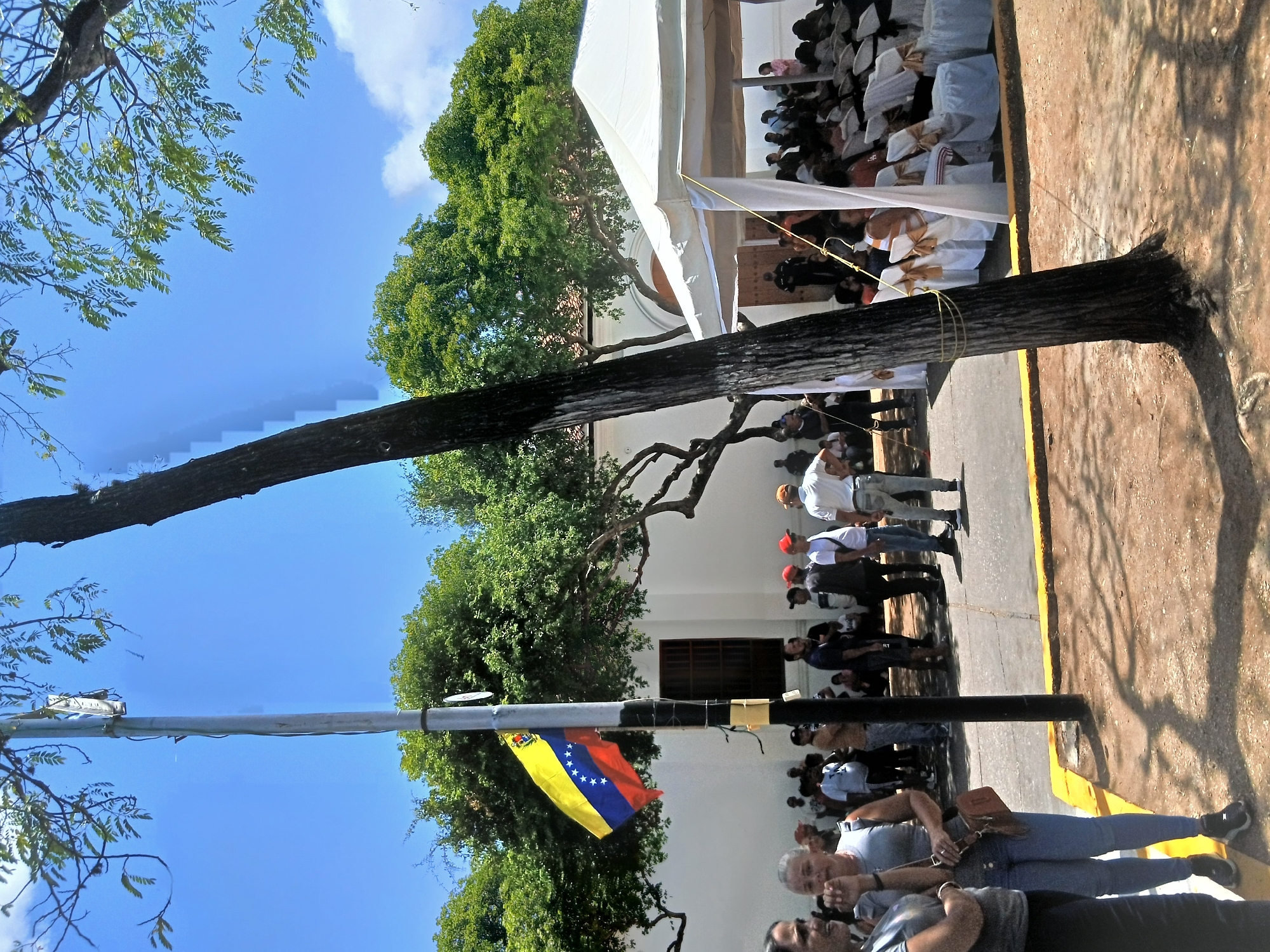 The image depicts a scene in Santa Ana, Venezuela. A crowd of people stands on a paved area next to a building with a white façade and a tent structure. A Venezuelan flag is attached to a pole in the foreground. A tree stands between the flag and the building. The sky is blue with some clouds, and trees are visible in the background.