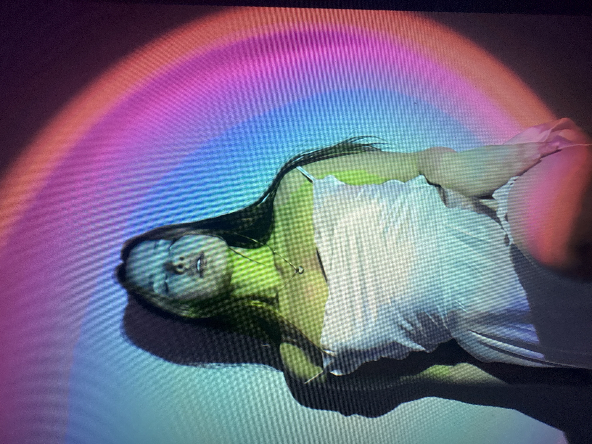 A woman lies on her side on a surface illuminated by a rainbow-colored light projection. She wears a white slip dress and a necklace. Her eyes are closed and her hair is dark. The location is undeterminable.