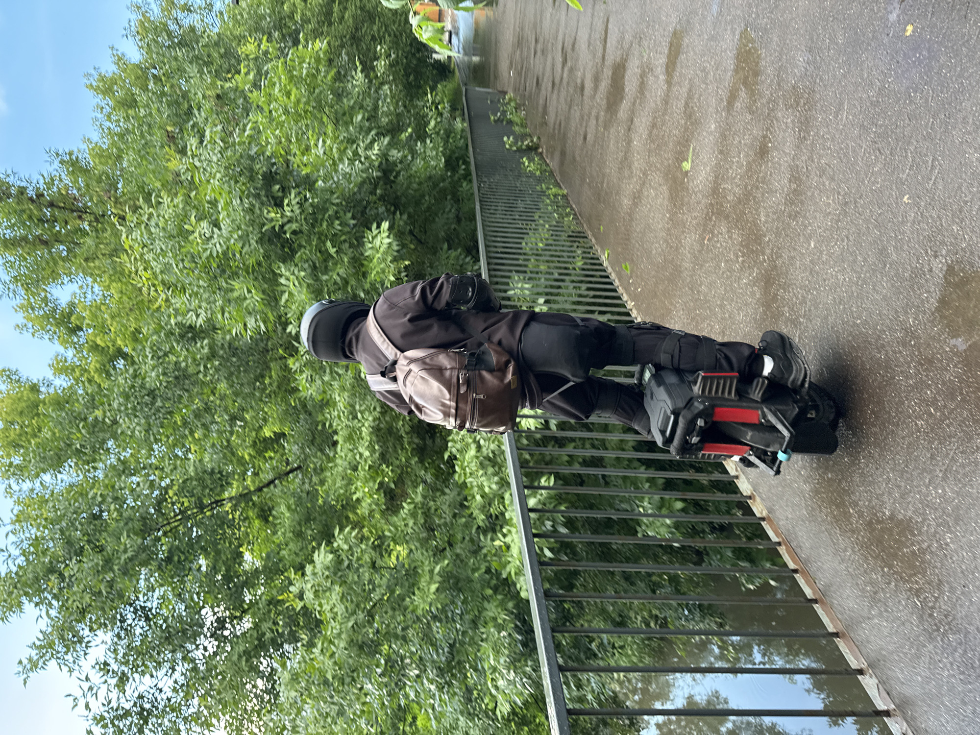 An individual wearing a black helmet, black jacket, black pants, and black shoes with white soles rides a black electric unicycle with red accents along a wet, paved pathway. The person carries a brown backpack. A dark metal railing runs along the left side of the pathway, bordering a body of water. Lush green trees and foliage are visible behind the railing and the pathway. The sky is blue with some clouds. The location is not identifiable as Moscow, Russia.