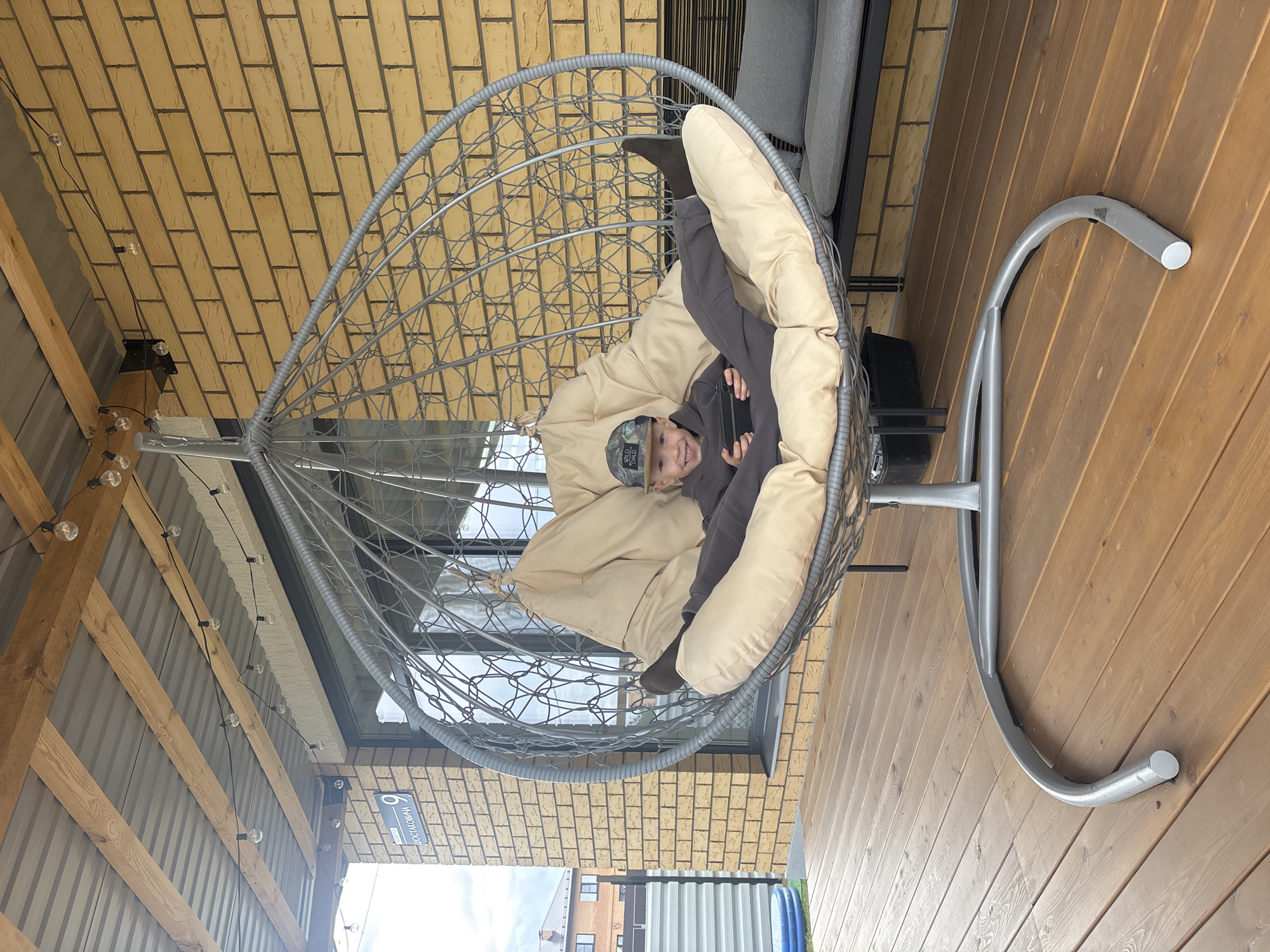A young child is comfortably seated in a large, grey woven hanging egg chair with soft, beige cushions. The child is wearing a dark grey tracksuit and a camouflage baseball cap, looking towards the viewer with a broad smile while holding a smartphone. Their legs are bent, and feet are tucked into the cushions.
The setting appears to be an outdoor patio or balcony. The flooring is made of wooden deck planks, and the structure behind the chair features a light brown brick wall and a section of corrugated metal siding with wooden support beams. String lights with round bulbs are strung along the wooden beams and under the eaves. Through a large window visible behind the chair, another building can be seen in the distance under what appears to be an overcast sky.
A grey metal stand supports the hanging chair, curving gracefully from the floor. On the corrugated metal wall, a blue and white street sign is visible, displaying the text "ЧАЙКОВСКОГО" (Chaykovskogo) and the number "9," indicating the location. The overall atmosphere suggests a relaxed daytime scene.