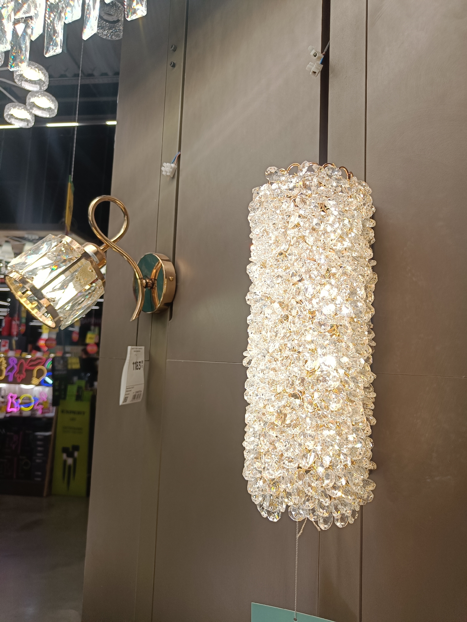 The image shows two wall-mounted light fixtures with a modern, decorative design, featuring faceted crystals. The left fixture has a single, angled crystal shade supported by a gold-toned curved arm. The right fixture is a vertical column covered in many small, clear crystals, also with gold-toned accents. Both are mounted on a plain, greyish-brown wall. In the background, there is a reflection of a ceiling light fixture composed of multiple faceted crystal elements. Below the left fixture, a price tag with the number "1183" is visible. Further in the background, out of focus, are shelves with various items, some of which appear to be illuminated neon signs in bright colors like pink, yellow, and blue, suggesting a retail or showroom environment. Electrical wire connectors are visible on the wall near the top of the fixtures. The overall impression is a display of decorative lighting in what appears to be a store.