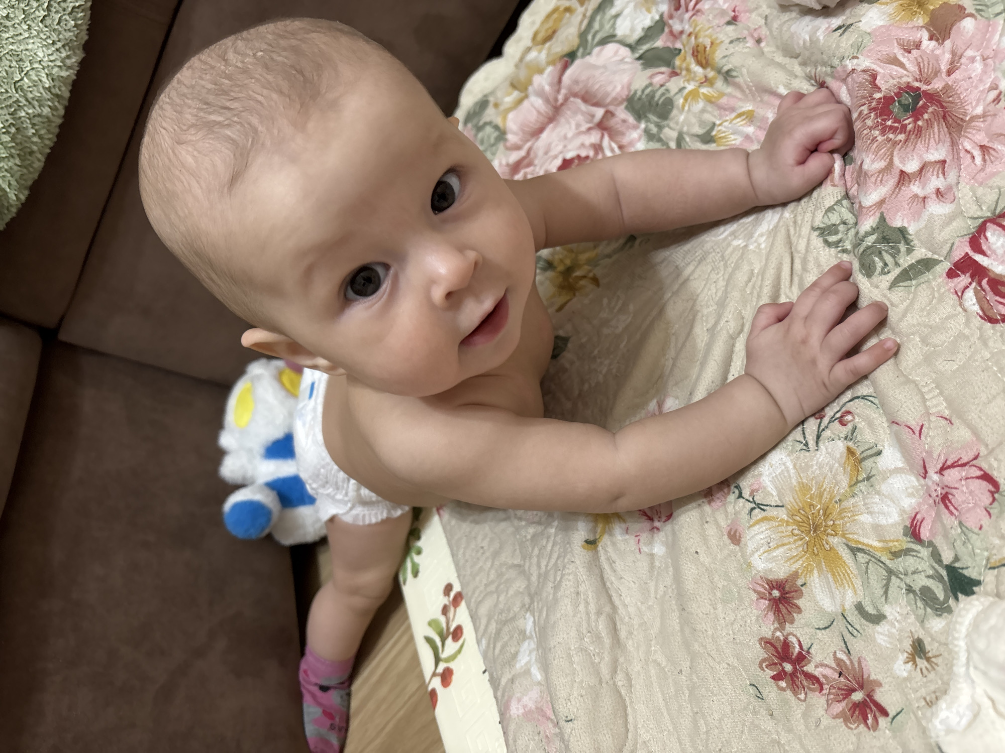 A baby with sparse hair, dressed in a diaper and pink socks, is positioned on a floral patterned quilt. The baby is looking up at the camera. A plush toy is situated near the baby's legs. The setting appears to be indoors. There is no indication of the city of Sochi, Russia in the image.