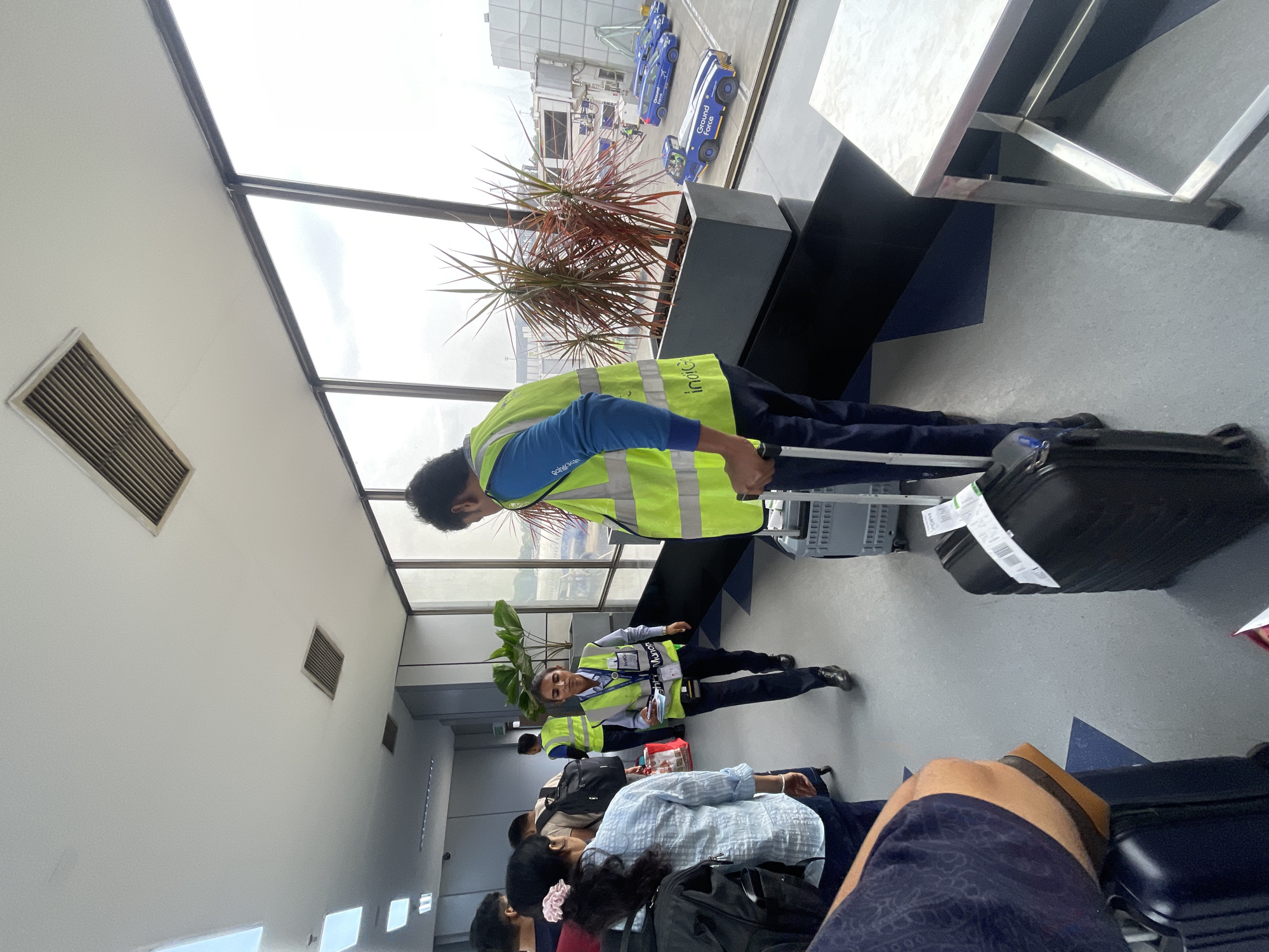 The image depicts an airport jet bridge in Ahmedabad, India, with passengers and airport staff. The environment features large windows overlooking an airfield with several blue vehicles, possibly ground support equipment, and airplanes. Inside the jet bridge, passengers are queuing with their luggage. Several individuals are wearing high-visibility vests, indicating they are airport personnel. One person in a bright yellow vest is interacting with a passenger, while another is seen walking with a suitcase. Plants in large planters are placed along the corridor. The floor has a geometric pattern of blue and grey tiles. The lighting suggests it is daytime.
