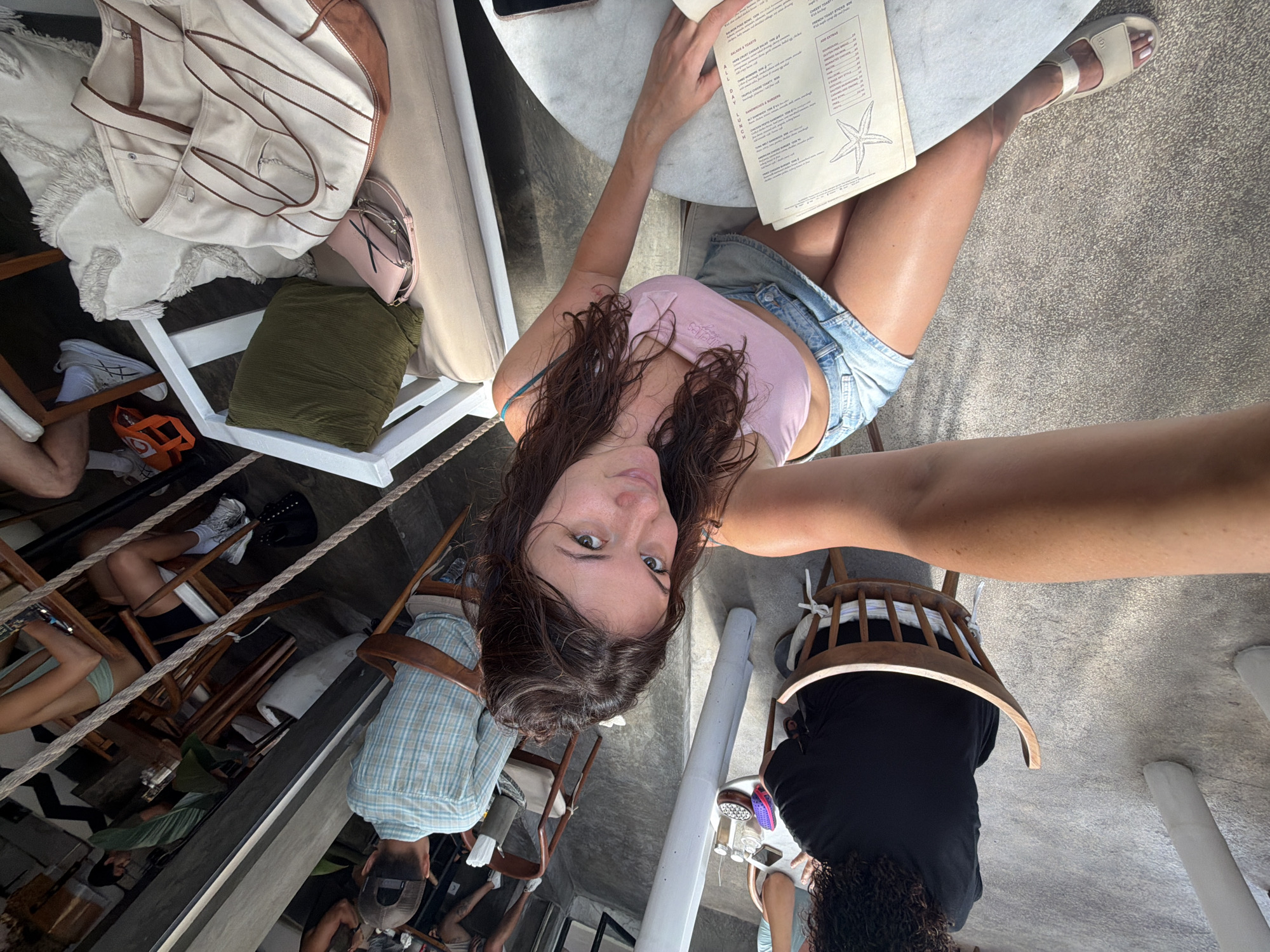 In North Kuta, Indonesia, a woman is seated at a table outdoors, holding a menu. She is wearing a pink crop top and denim shorts. Beside her, another person with curly hair is wearing a black shirt. The scene takes place in a cafe or restaurant with wooden chairs and tables. In the background, other patrons are visible, some seated and some standing, with various furniture and decorative elements. The overall atmosphere appears casual and relaxed, with natural light filtering into the space.