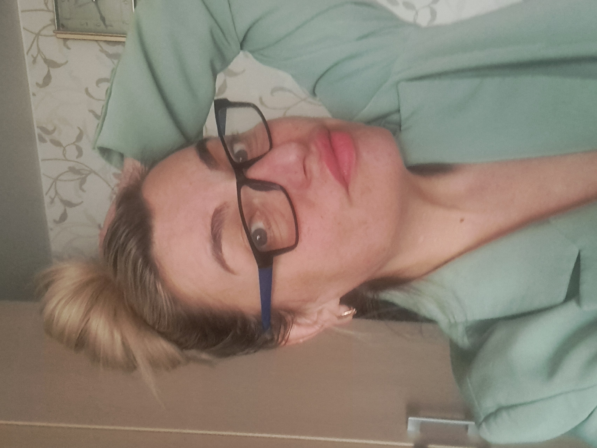 A woman with blonde hair in a bun, wearing black-framed glasses and a light green jacket, is lying on her back. A picture frame with a clock is visible behind her. The photograph appears to be taken indoors and is not definitively tied to Elektrostal, Russia based solely on the visual information available.