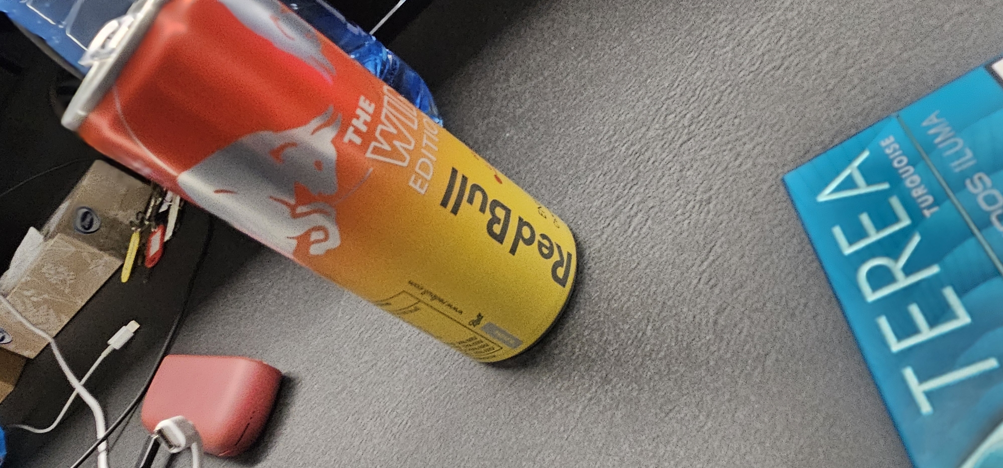 The image shows a partially visible can of Red Bull The White Edition, tilted to the left, with its red and silver top and yellow body. To the right of the can is a blue box labeled "TEREA TURQUOISE," "IDOS ILUMA," and additional text.
To the left of the can is a set of keys, a small cardboard box, a white cable, and a small pink object. The background is a neutral gray surface. The location is unknown and there is no way to know it is in the city of Sofia, Bulgaria. There are no people present, and no actions or interactions depicted.