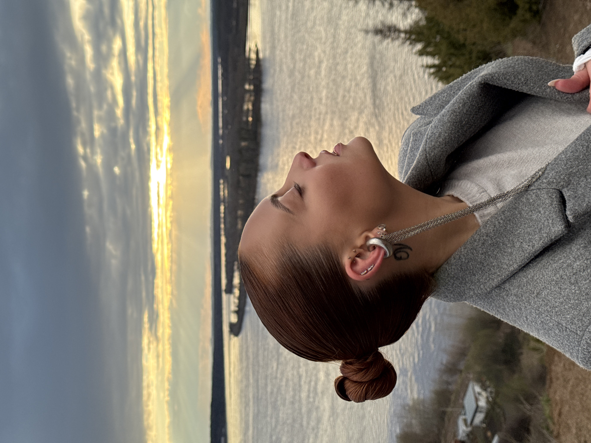 The image depicts a tranquil outdoor moment at what appears to be sunset or sunrise, with a vast body of water and a dramatic sky forming the backdrop. The subject, a person seen in profile, gazes upwards with eyes closed or nearly closed, their expression serene and contemplative. Their reddish-brown hair is pulled back into a tight, neat bun. They are dressed in a light grey top and a charcoal grey jacket or coat, adorned with a silver chain necklace.
Notable details include multiple ear piercings on the visible left ear, featuring a silver tunnel in the stretched lobe, a curved barbell piercing the helix, and an industrial bar. A small, stylized black tattoo, possibly resembling a 'G', is visible behind the ear. The setting appears to be a natural landscape, likely in Ольховка, Russia, overlooking a large lake or river. The sky is a prominent feature, showcasing vibrant golden light from the sun piercing through scattered clouds, creating striking sunbeams and a warm glow across the water. The distant shoreline is lined with faint trees and structures, adding depth to the expansive scene. The overall atmosphere conveys peace and appreciation for the natural beauty.