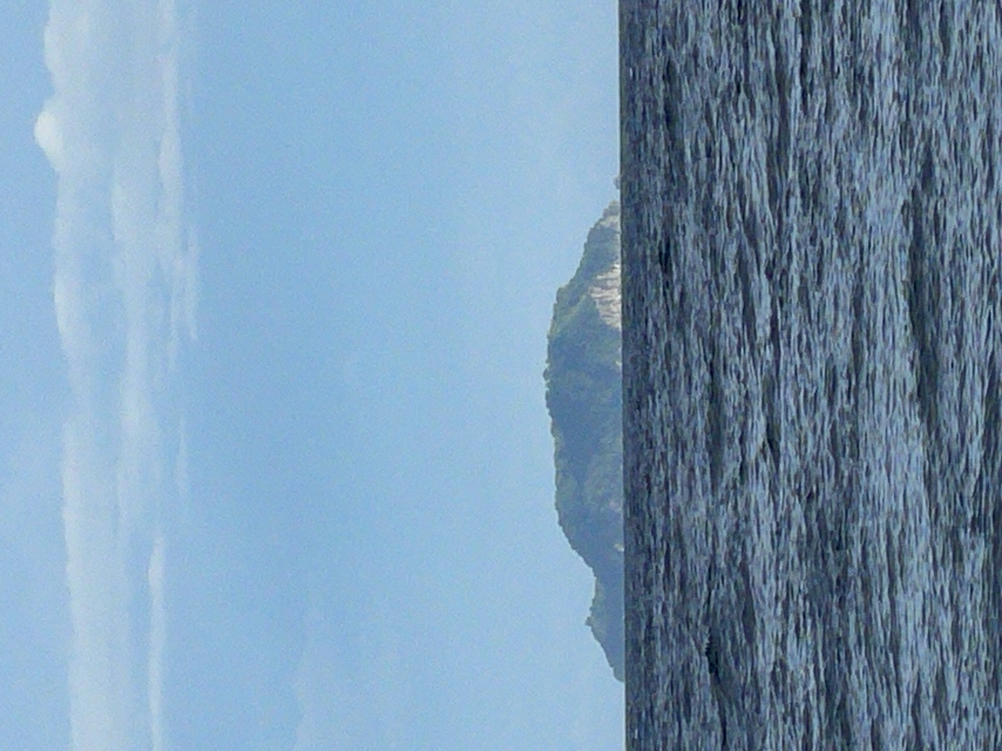 A wide-angle, eye-level shot captures a distant, green, mountainous island situated in the ocean. The island's terrain is rugged and appears covered in dense vegetation. A visible cliff face is present on the island's side. The ocean in the foreground is dark blue and slightly choppy, with ripples indicative of a gentle breeze. The sky above is a clear, light blue, with a few wispy white clouds scattered across the horizon. The scene suggests a natural, undisturbed landscape. This island is likely Apo Island, a protected marine sanctuary located off the coast of Dauin, Negros Oriental, Philippines.