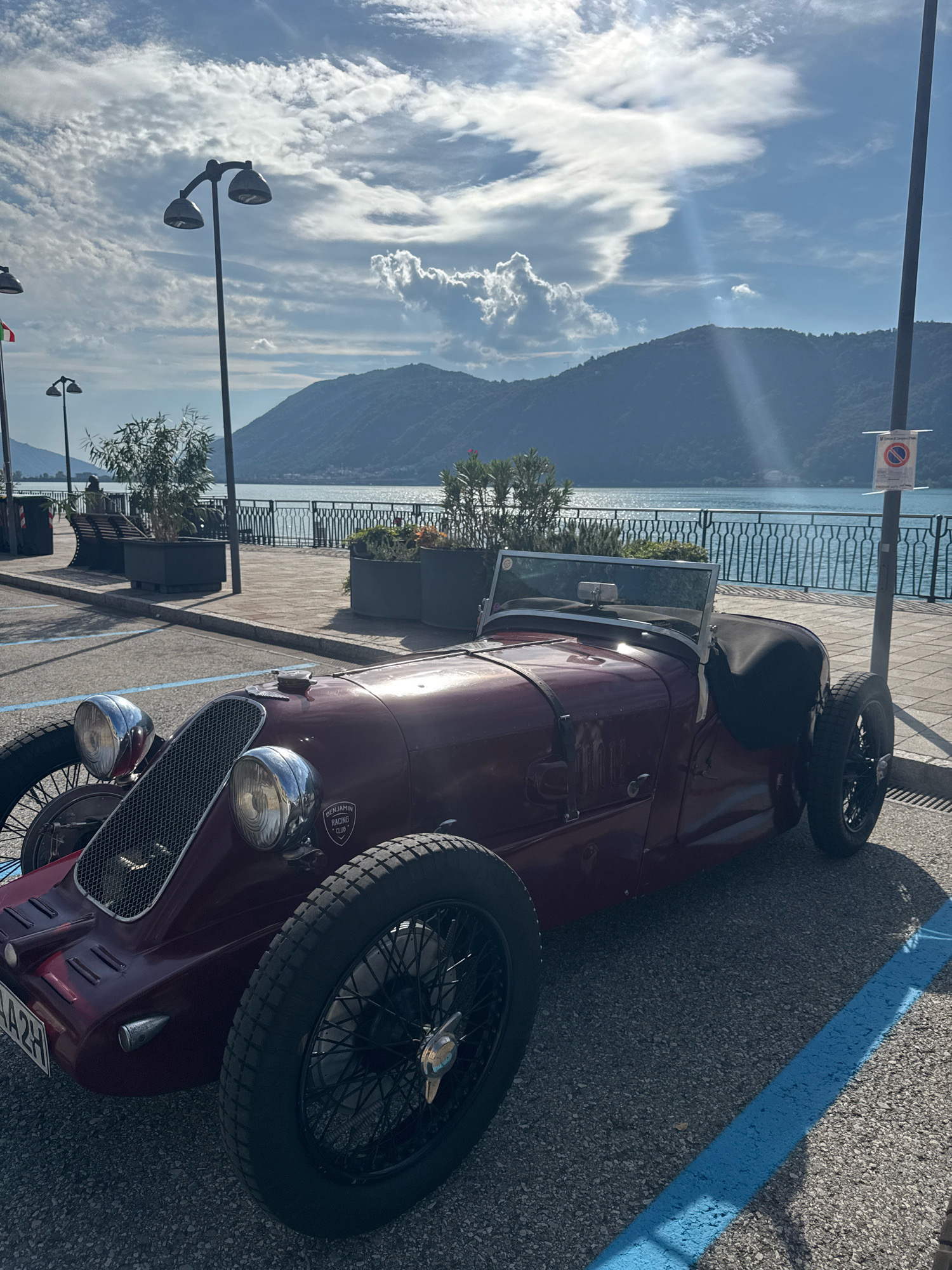 The image depicts a vintage, maroon-colored convertible sports car parked on a paved surface in Campione d'Italia, Italy. The car is positioned at an angle, with its front facing towards the left side of the frame. Its design features prominent, circular headlights, a distinctive grille, and exposed wire-spoke wheels. The car's hood has several straps, and a small windshield is visible. A black fabric cover is draped over the passenger side of the car.
In the background, a body of water, likely Lake Lugano, is visible, with lush green hills rising on the opposite shore. A metal railing separates the car's parking area from a promenade or walkway that runs along the waterfront. Several lampposts, benches, and planters with vegetation are present along this walkway, suggesting a public space. The sky is partly cloudy, with shafts of sunlight breaking through, casting shadows on the ground. A parking sign with a red circle and a diagonal line is affixed to a pole on the right side of the frame, indicating a prohibition, possibly related to parking. The overall scene suggests a sunny day in a scenic Italian town.