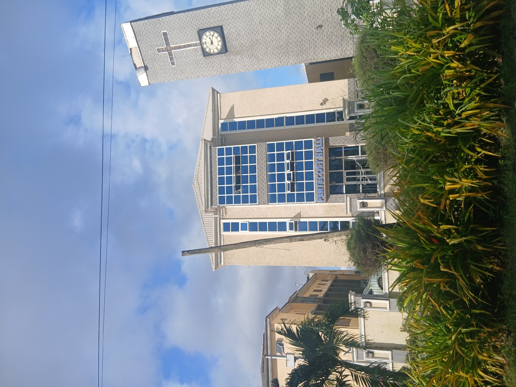 The image shows the Pentecost Hall, a modern building with a beige facade and blue glass windows, located in Isunba, Nigeria. To the right of the hall is a tall, grey bell tower with a cross and a clock face. The clock shows approximately 3:30. In front of the hall, lush green and yellow foliage is visible, with a palm tree to the left. The sky is a clear blue with scattered white clouds. A single power line stretches across the upper portion of the image.