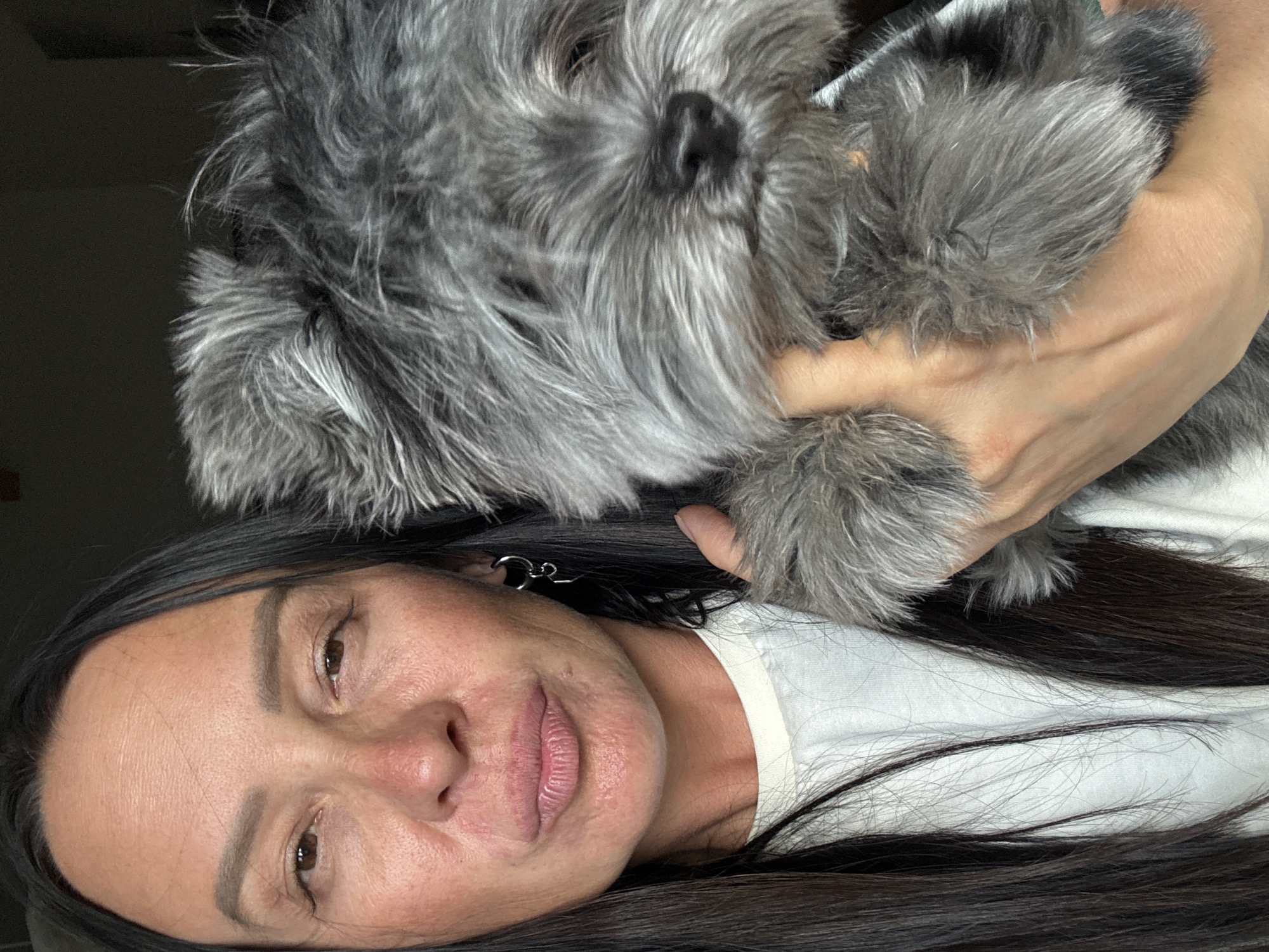 A woman with long, dark hair is holding a gray, fluffy dog in her arms. She is wearing a white t-shirt and silver earrings. The background is dark and nondescript. The location is not discernible from the image.