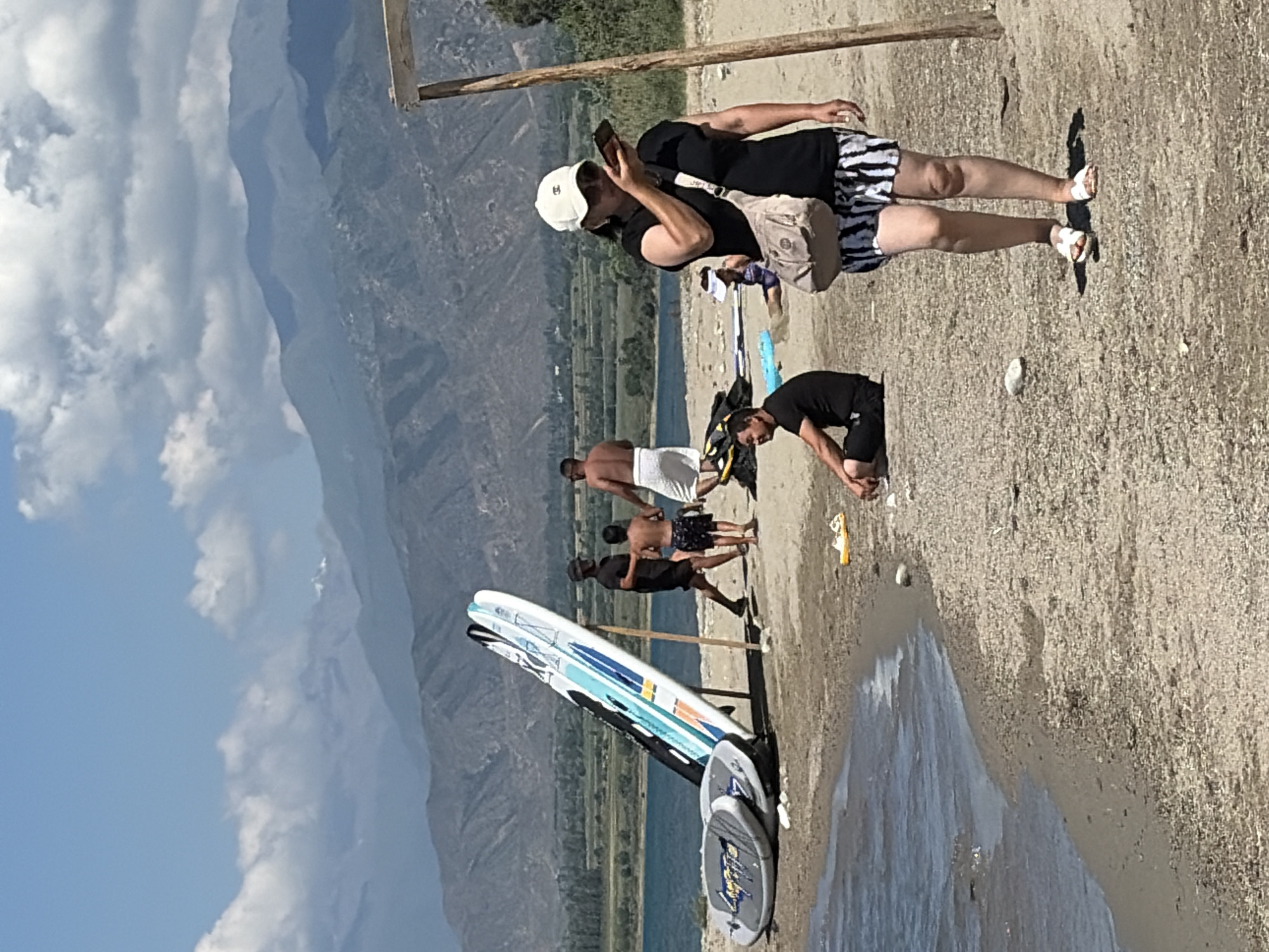 A beach scene unfolds with a body of water in the foreground, bordered by a sandy shore. Several paddleboards are propped up, leaning against a wooden support structure. In the middle ground, a group of people are visible: two men standing, one with a towel wrapped around his waist, and a child wearing swim trunks. Another man is seated on the sand. In the foreground, a woman wearing a black t-shirt, a white baseball cap, and patterned shorts is looking at her phone. She carries a beige shoulder bag and is wearing white sandals. The background features a vast expanse of mountains under a partly cloudy sky. The overall environment suggests a recreational area at a lake or reservoir.