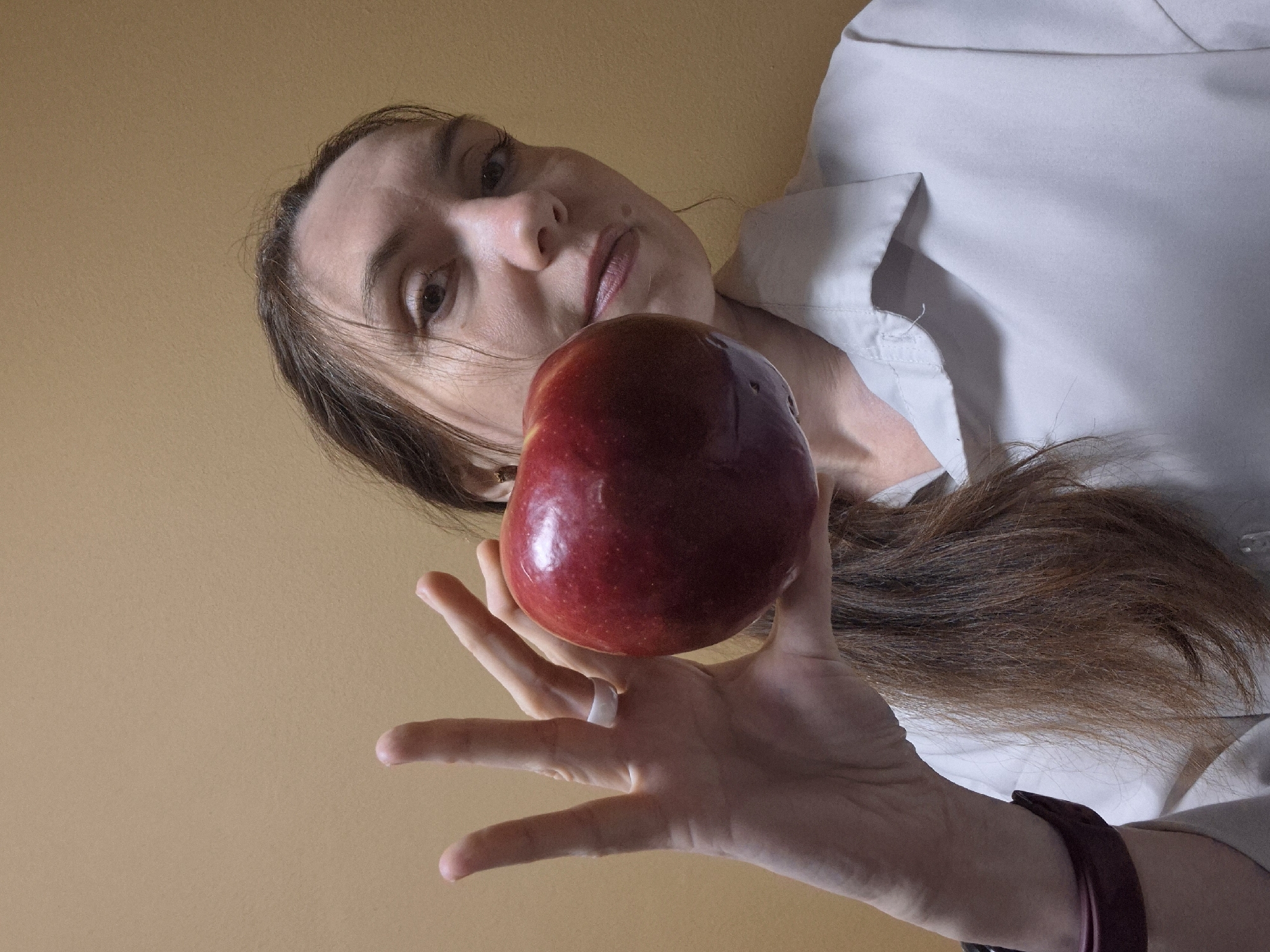 A low-angle, close-up shot captures a woman holding a red apple directly above the camera. The woman, with long, dark brown hair, looks downward with a neutral to slightly serious expression. Her eyes are open and gaze directly at the viewer. She is wearing a light-colored, possibly white or very light grey, collared shirt.
Her right hand (from the viewer's perspective) extends into the frame from the lower right, holding the bright red apple. A white ring is visible on her ring finger. A dark, possibly leather, wristband or bracelet is on her wrist, partially obscured by her hair. The apple itself is prominently featured, appearing fresh with a slightly irregular shape and a small dark indentation on its surface.
The background is a plain, light brown or tan wall, suggesting an indoor setting. The lighting is soft and even, without harsh shadows, and the time of day is not discernible. No text is visible in the image.
