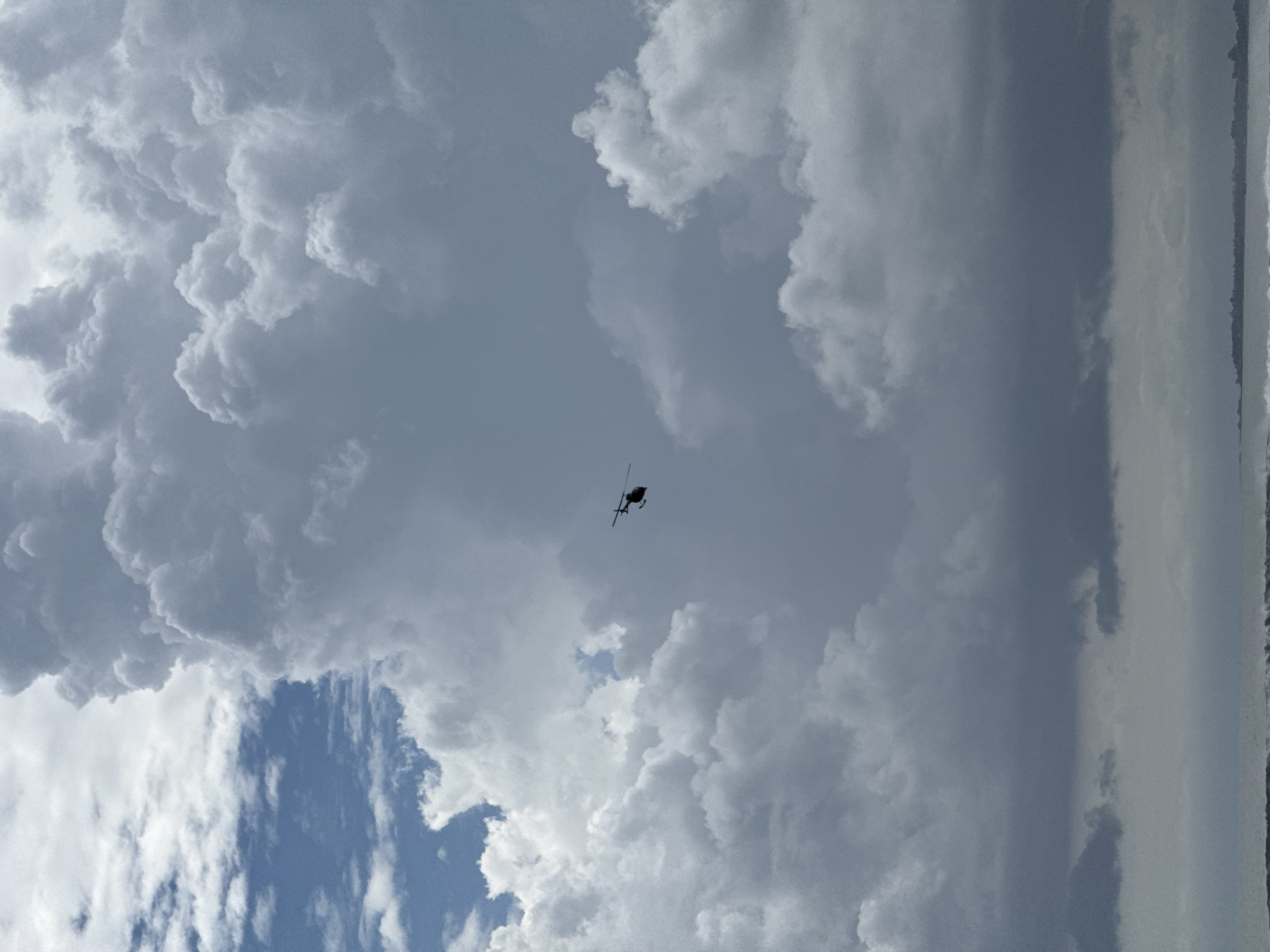 The image depicts a helicopter flying in the sky. The sky is filled with dark, ominous cumulonimbus clouds. To the side of the image is what could be the ocean horizon. Kuta, Indonesia is not apparent in the image.