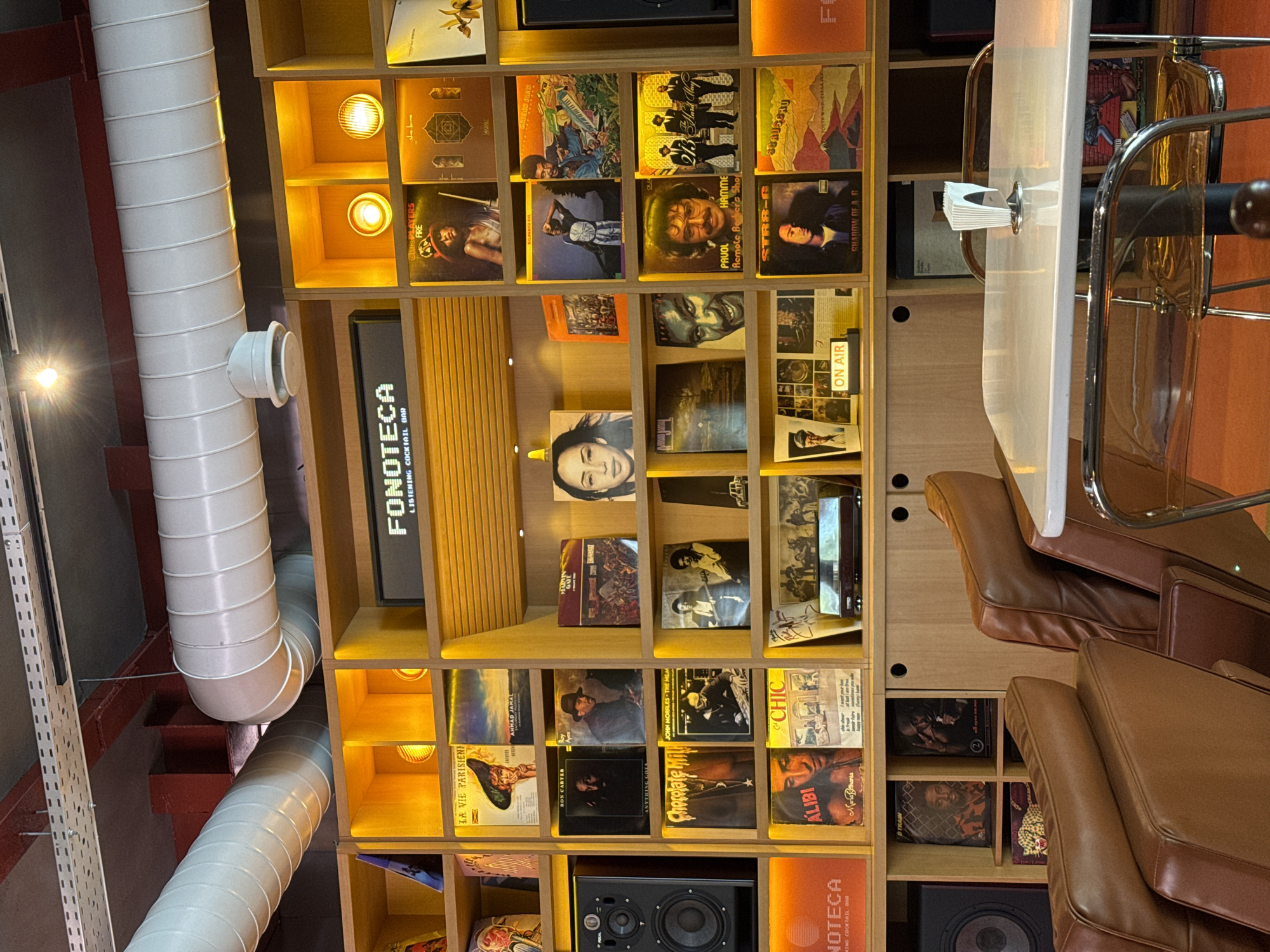 The image displays the interior of a modern listening cocktail bar, likely located in Yerevan, Armenia, characterized by a blend of cozy and industrial design. A prominent feature is a large, custom-built wooden shelving unit filled with numerous vinyl record albums displayed facing forward, each cubby illuminated by warm, recessed spotlights.
To the right, a white table with a napkin holder is accompanied by comfortable brown leather armchairs, suggesting a relaxed seating area for patrons. The left side and ceiling feature large, white corrugated ventilation pipes and exposed metal conduits, contributing to an industrial aesthetic. Various light sources, including an exposed bulb, provide warm illumination throughout the space. Black audio speakers are visible on the lower shelves, indicating the bar's musical focus.
A vertical black sign with white lettering clearly states "FONOTECA LISTENING COCKTAIL BAR," while a smaller sign reads "ON AIR," possibly indicating a live session or current broadcast. Album titles and artists visible on the shelves include "LA VIE PARISIENNE," "BON CARTER" (Ron Carter), "ANYTHING GOES," "Chocolate Milk," "AHMAD JAMAL," "JOHN MORALES THE M+M," "ALIBI," "CHIC," "The Ohio Players FIRE," "GILBERTO GIL," "PAUL HAMMER" (Paul Hardcastle), "STR8-G," and "SHADOW OF A D.B." The overall setting evokes a warm, inviting atmosphere designed for music enthusiasts to enjoy cocktails and records.