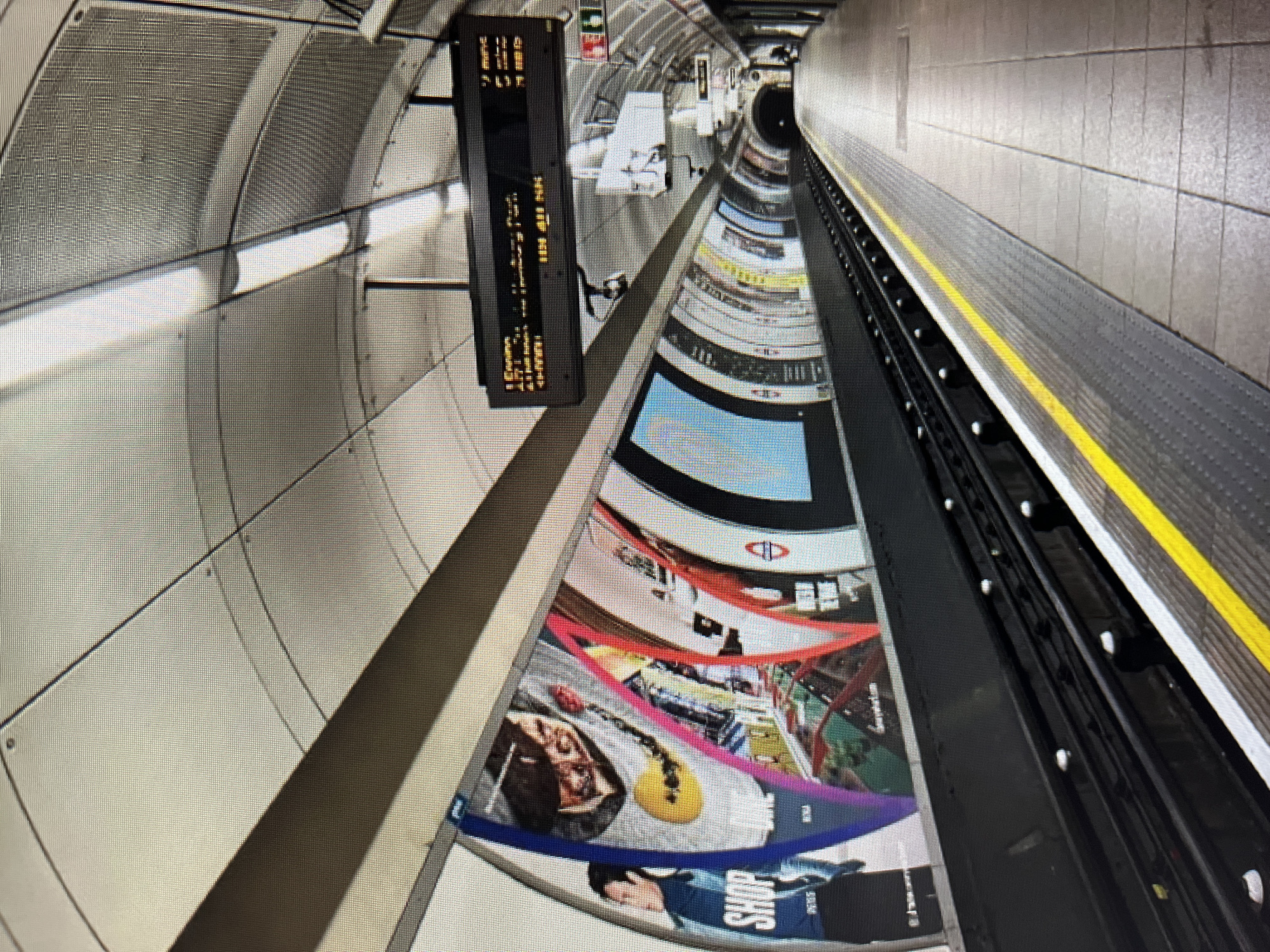 This media file depicts an interior view of a subway or train station tunnel. The scene is dominated by the curved architecture of the tunnel walls, which are covered in a large, curved advertisement. The advertisement features a collage of images, including food items like pizza, a yellow guitar, and what appears to be a shopping scene with people in the foreground. Text visible on the advertisement includes "SHOP".
On the left side of the image, a vertical digital display shows some information in a yellow font against a black background. The text is difficult to read clearly but appears to be time-based information, possibly train arrival times or station information. Above this display, the wall has a textured, metallic, grated appearance.
To the right of the tunnel, a platform edge is visible with a yellow safety line. Beyond the platform, the train tracks are visible, and further down the tunnel, another platform edge can be seen. The lighting appears to be artificial, emanating from lights running along the ceiling of the tunnel. The perspective is wide-angle, emphasizing the curvature of the tunnel and the advertisement. The overall impression is that of a modern, busy underground transportation hub. No specific time of day or weather conditions are discernible.