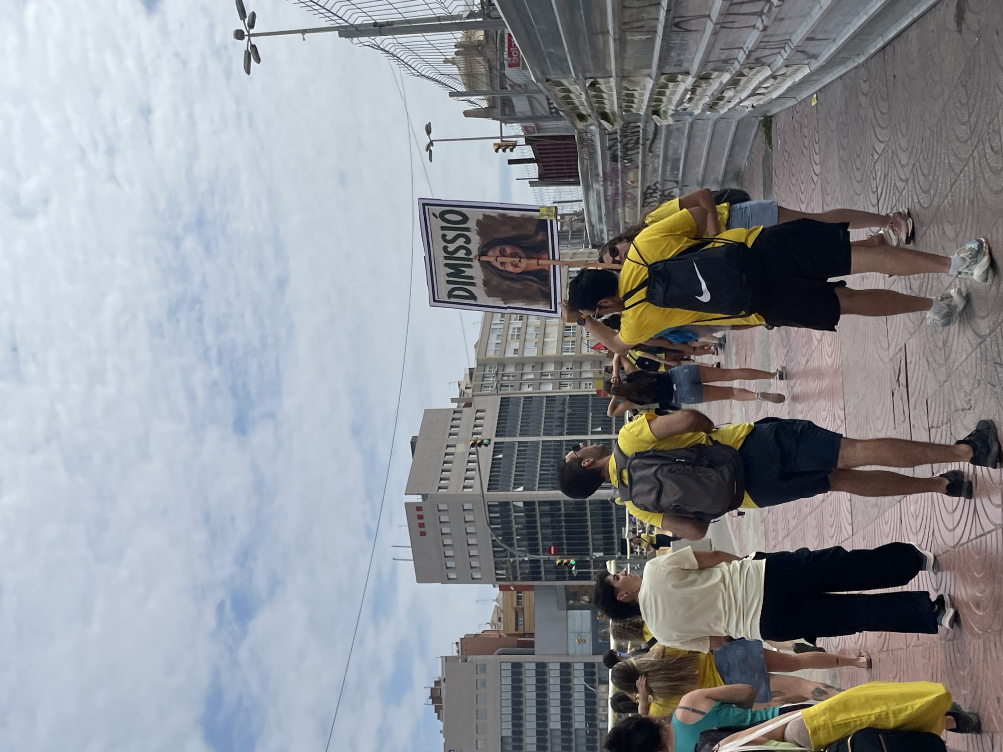 The image depicts a street scene in Barcelona, Spain, with a group of people walking along a sidewalk. In the foreground, two men in yellow shirts and black shorts are visible, both wearing backpacks. One man is holding up a sign with a picture of a woman and the word "DIMISSIÓ" in large letters. The background shows several multi-story buildings, street lights, and construction fencing. The sky is cloudy. The overall impression is that of a demonstration or protest.
