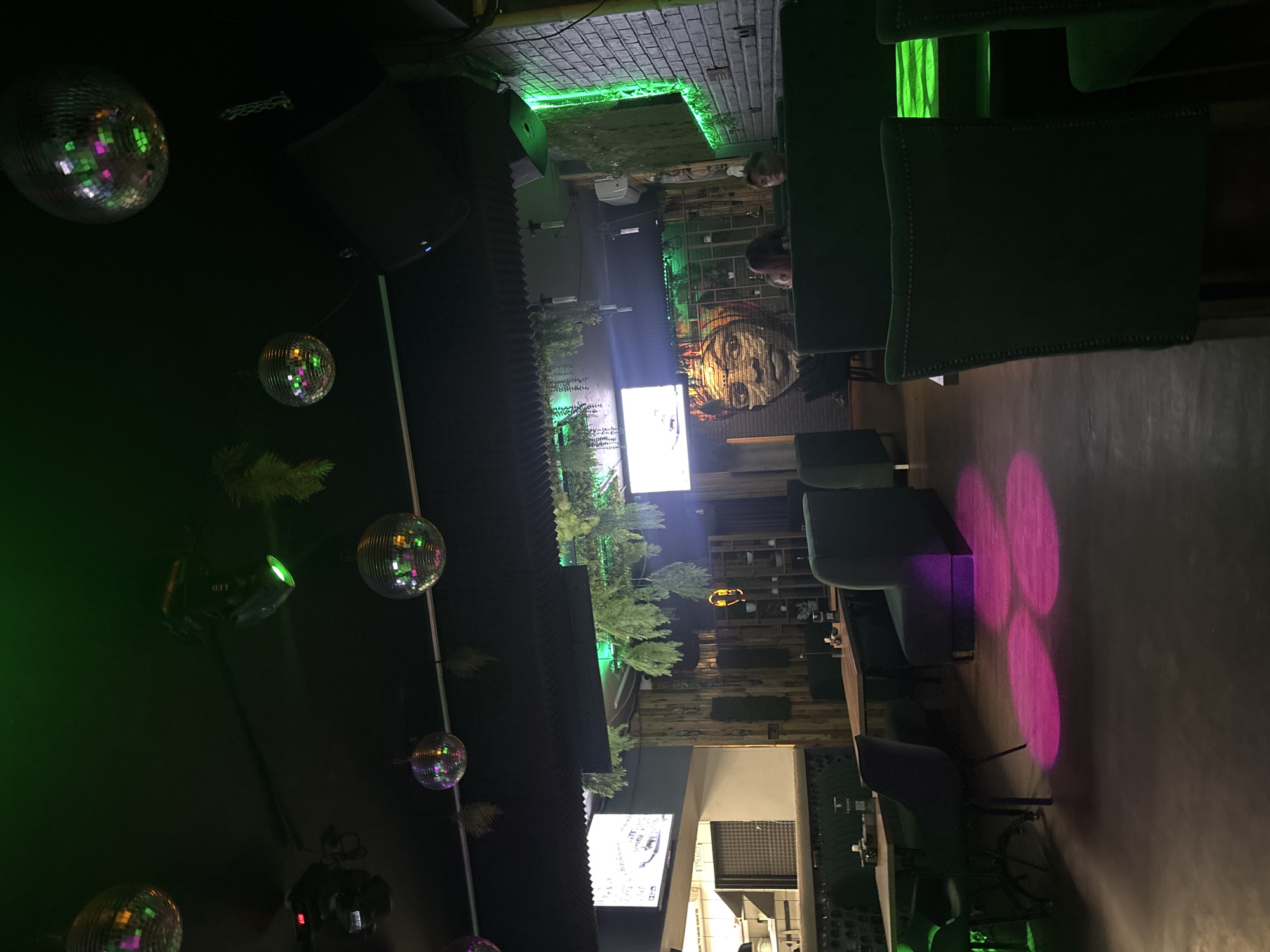 The image depicts the interior of a dimly lit bar or lounge, likely a shisha lounge, given the general ambiance and decor. The environment features dark walls and ceilings, punctuated by vibrant green and purple lighting. Several disco balls are suspended from the ceiling, reflecting the ambient light. Green foliage hangs from the ceiling and walls, adding a touch of natural element to the artificial lighting.
In the foreground, plush dark green seating, likely booths and chairs, are arranged around tables. The floor is dark and appears to be a polished concrete or similar material, with circular pink light projections scattered across it.
In the midground, more tables and seating are visible, along with a wall adorned with a large mural of a woman's face. A few patrons are seated in the background, with their faces partially obscured. One screen is visible on the left wall, displaying what appears to be a news broadcast or digital signage. On the right wall, exposed brick is visible, illuminated by bright green neon lighting.
The overall impression is a modern, trendy establishment designed for a nightlife atmosphere, with a focus on mood lighting and eclectic decor.