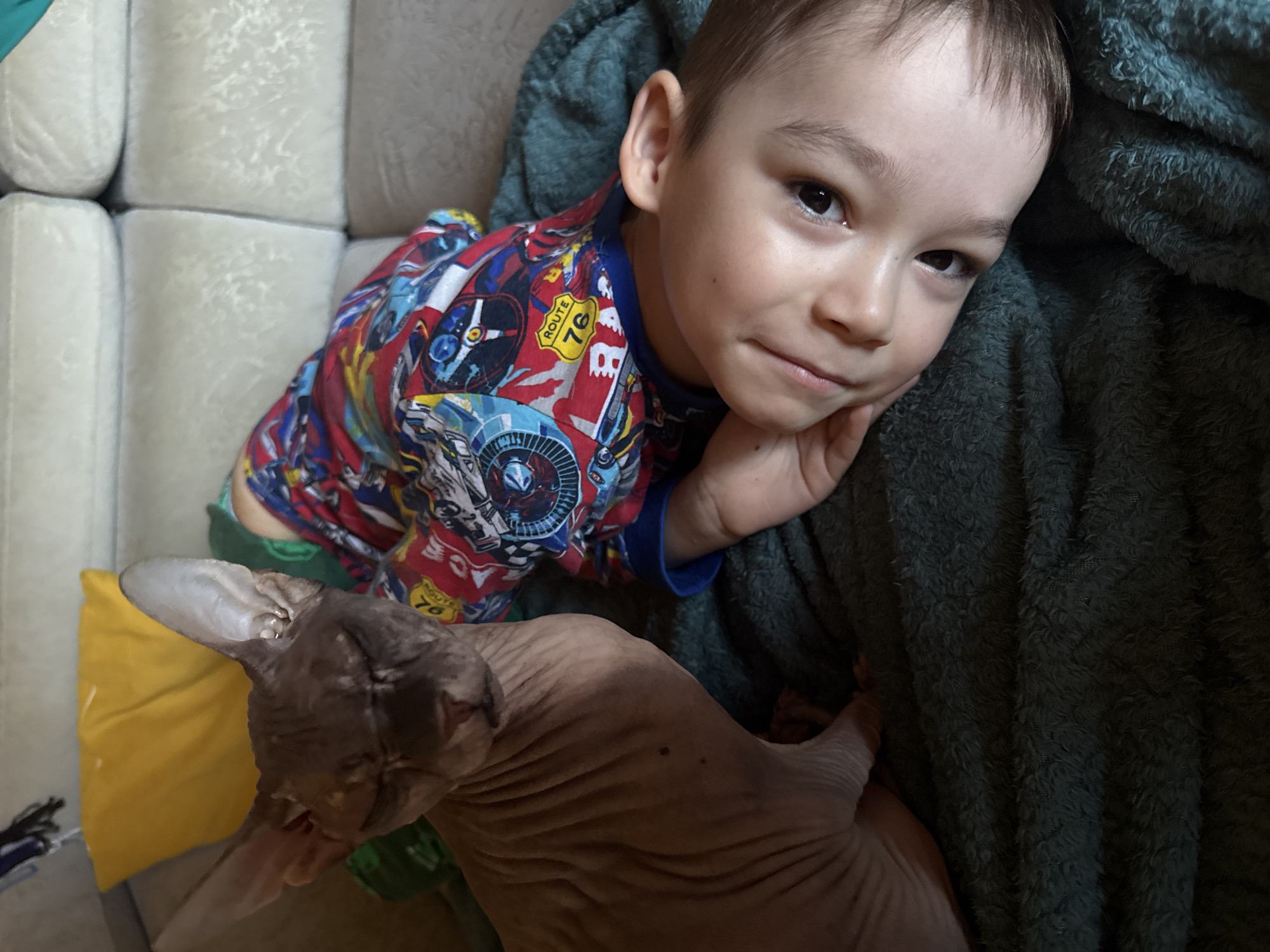 A Sphynx cat sits on a dark green fuzzy blanket to the left of a young child lying on their side on a light-colored couch. The cat is facing forward with its eyes closed and its ears perked up. The child is wearing a colorful shirt with a race car theme and is looking towards the camera with a slight smile. The child's head is propped up on their hand. A yellow pillow is visible in the background behind the cat. The scene appears to be indoors, likely in a living room.