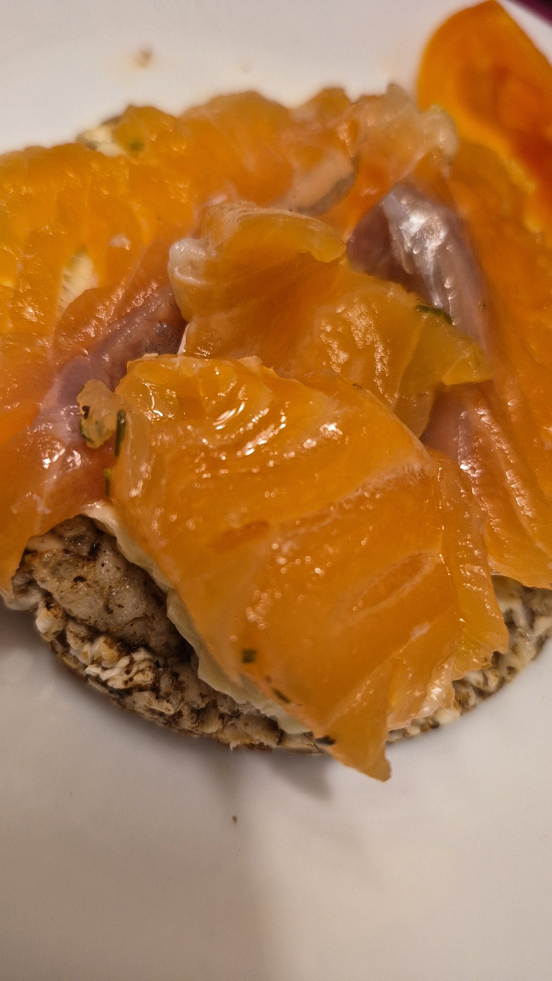 A close-up, overhead shot shows a single rice cake topped with cream cheese and thin slices of smoked salmon. The salmon is bright orange with some white streaks and appears slightly glistening. The rice cake has a dark, textured surface. The background is a plain white surface. There are no people or other objects visible in the frame. The scene does not provide any information that would allow for location in Krauja, Latvia.