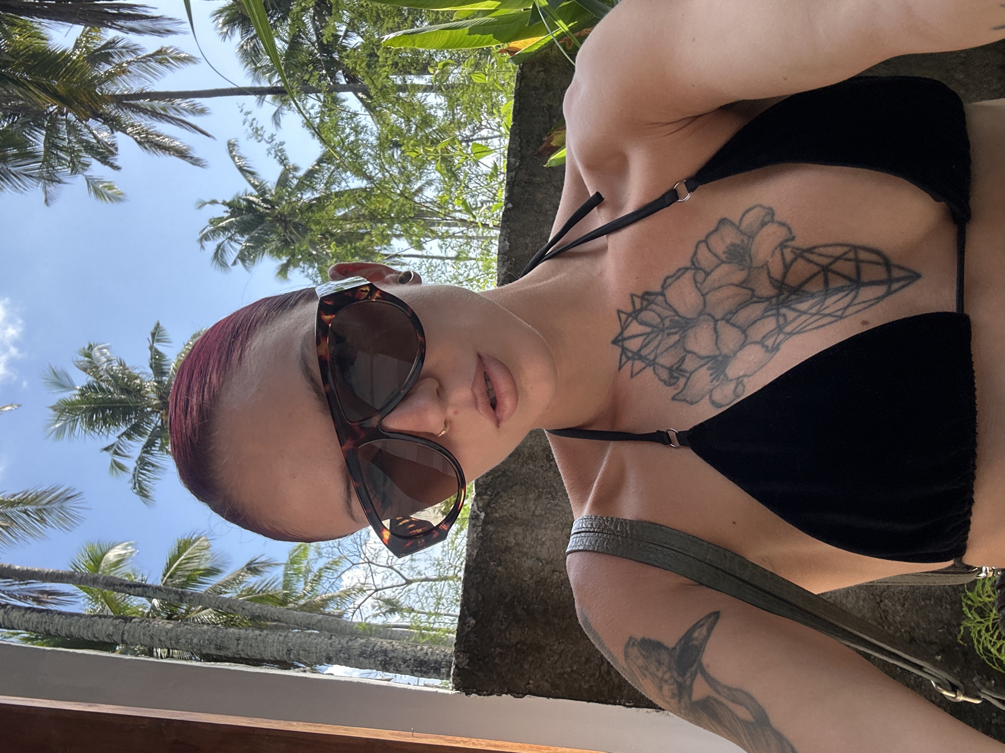 A person with red hair and sunglasses is wearing a black bikini top. They have tattoos on their chest and arm. Palm trees are visible in the background, indicating a tropical location. The photo was likely taken in Nusa Penida, Indonesia, based on the prevalence of palm trees and tropical vegetation. The person appears to be taking a selfie.