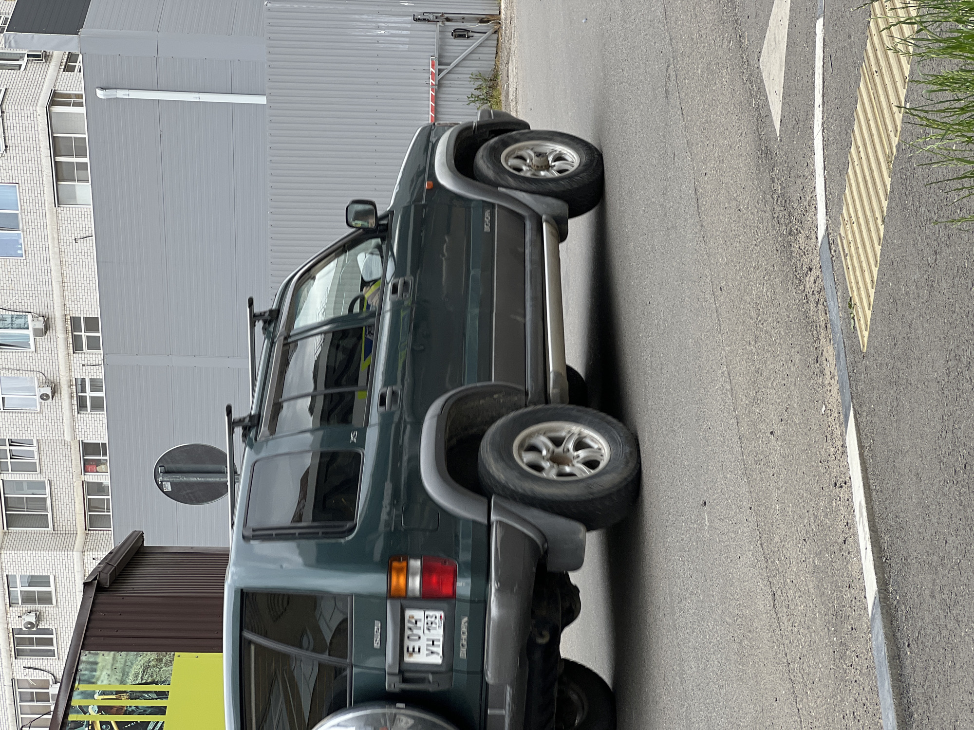 The image shows a dark green Isuzu Bighorn SUV with a spare tire mounted on the rear, driving on a paved road in an urban environment. The license plate reads "E 014 YH 193". The vehicle is captured in motion, with a slight blur. In the background, there is a light brick building with multiple windows, a corrugated metal fence, and a section of a gray building with a white downspout. A red and white striped barrier arm is visible on the right side of the image. The foreground features a yellow tactile paving strip and some green grass. The scene appears to be located in an industrial or commercial area.