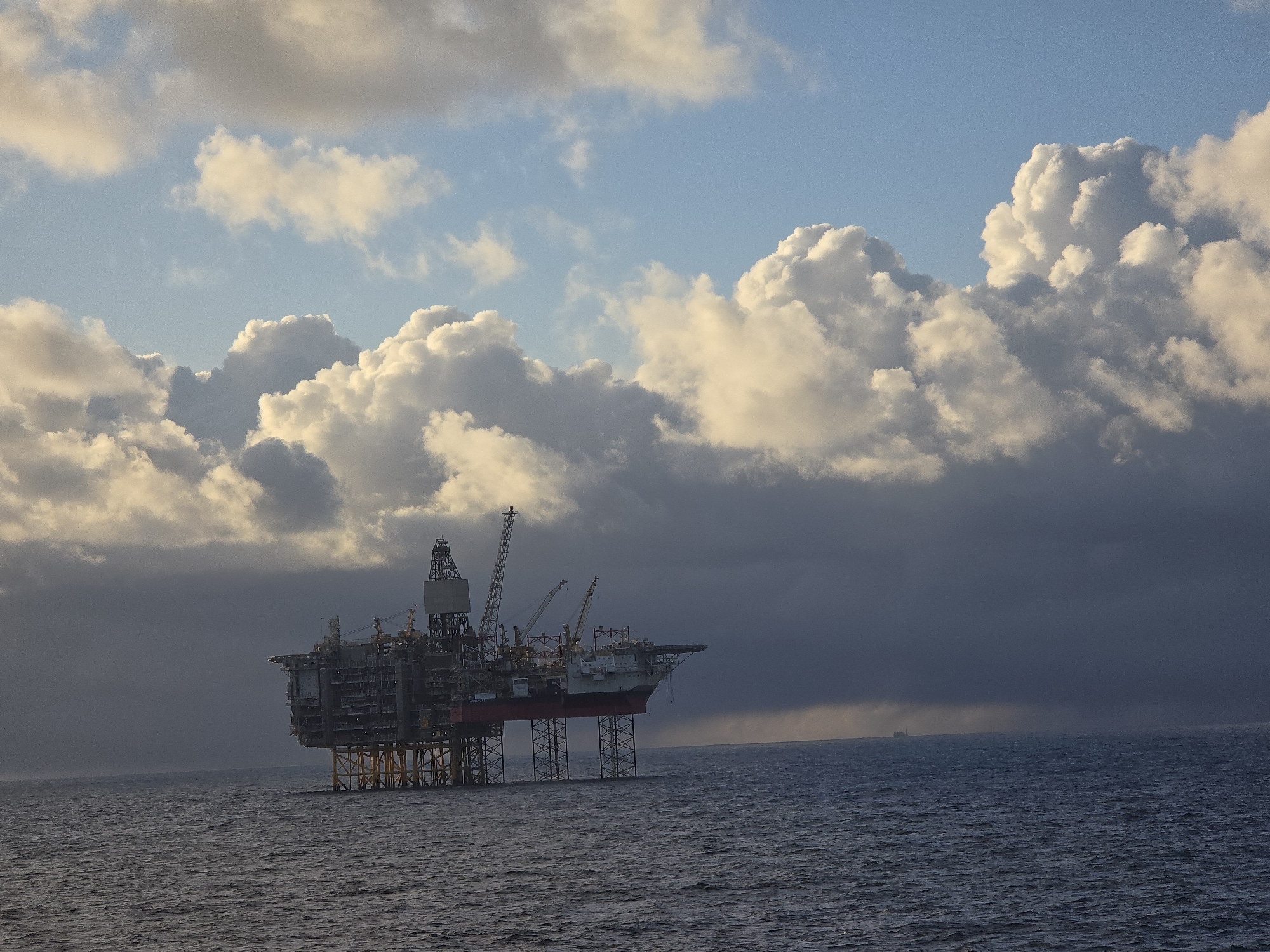 The image shows an offshore oil rig in an ocean environment. The rig is a complex structure with multiple platforms, cranes, and support legs extending into the water. The sky is filled with dramatic clouds, ranging from light and fluffy to dark and ominous, suggesting an approaching storm. The ocean is choppy with visible waves. A small, indistinct ship is visible in the distance.