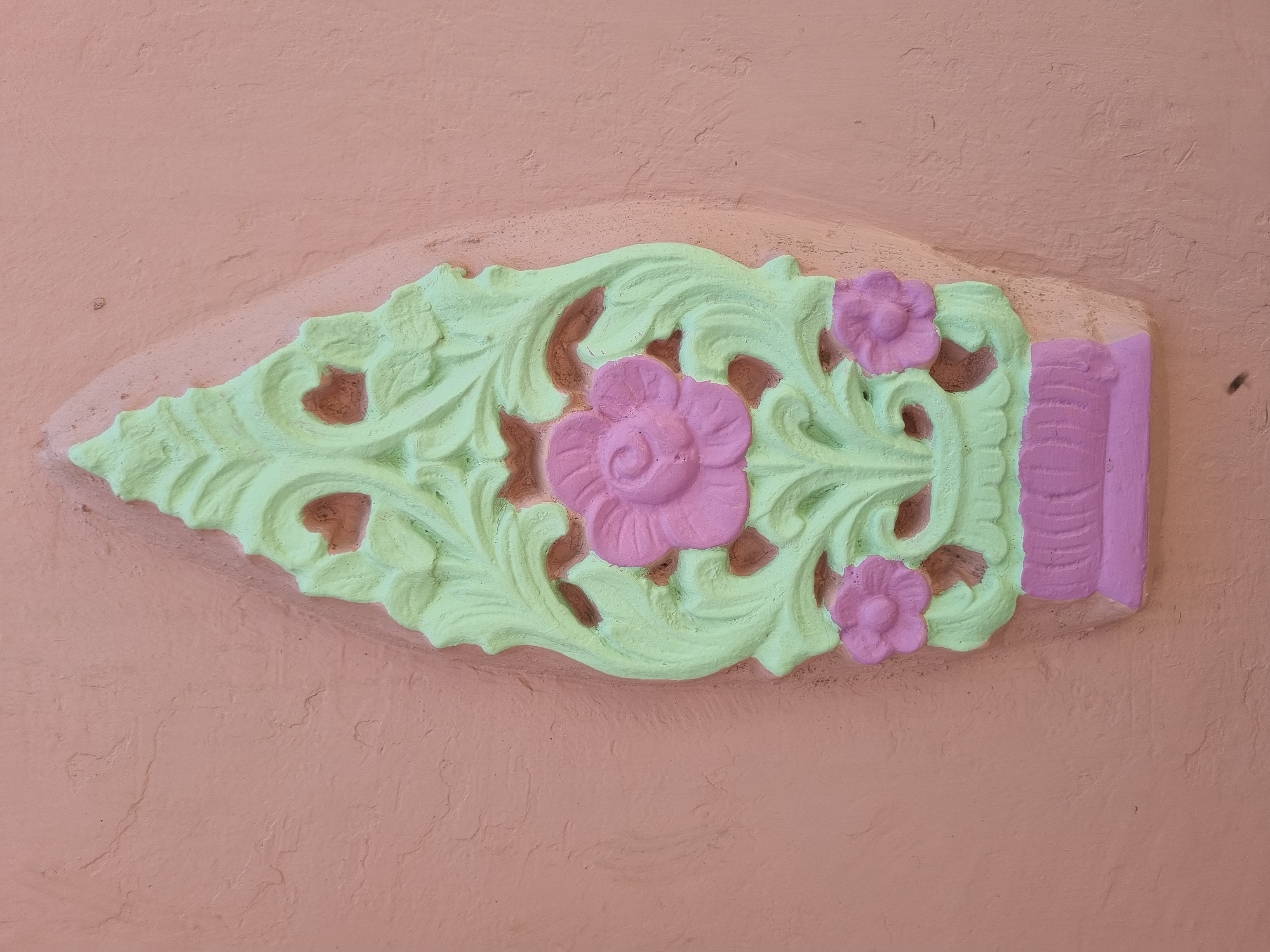 A vertically oriented, sculpted relief is centrally affixed to a textured peach-colored wall in Pedda Kammavaripalli, India. The ornamental relief, approximately flame-shaped, features stylized floral and scrollwork motifs. Its primary color is pastel green, highlighting detailed leaf-like forms. A large, pink, multi-petal flower is positioned centrally, with two smaller pink flowers situated near the bottom. A solid purple, ribbed, vessel-like base supports the green structure. The surrounding wall surface exhibits minor irregularities and a matte finish. No people, actions, or other objects are visible.