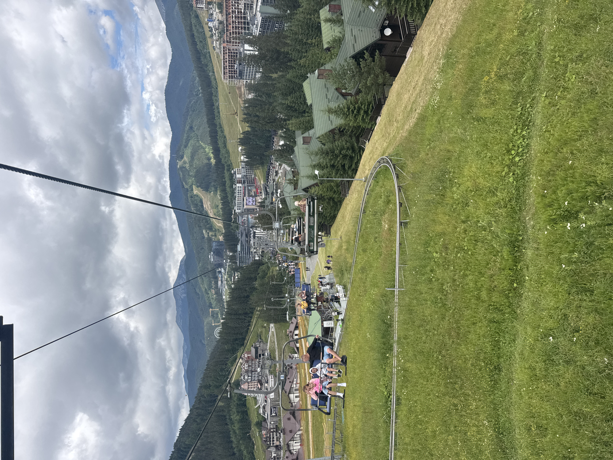 The image captures a scenic view from a ski lift in Polianytsia, Ukraine. In the foreground, a ski lift carries four individuals. Two adults and two children are seated, with the children appearing to be young. The ski lift is moving along a cable suspended over a grassy slope. In the middle ground, a village with traditional architecture and green-roofed houses is nestled amongst evergreen trees. Beyond the village, a cluster of modern hotels and residential buildings are visible. The background reveals a lush, forested mountain range under a cloudy sky. The overall scene suggests a summer day in a mountainous resort town.