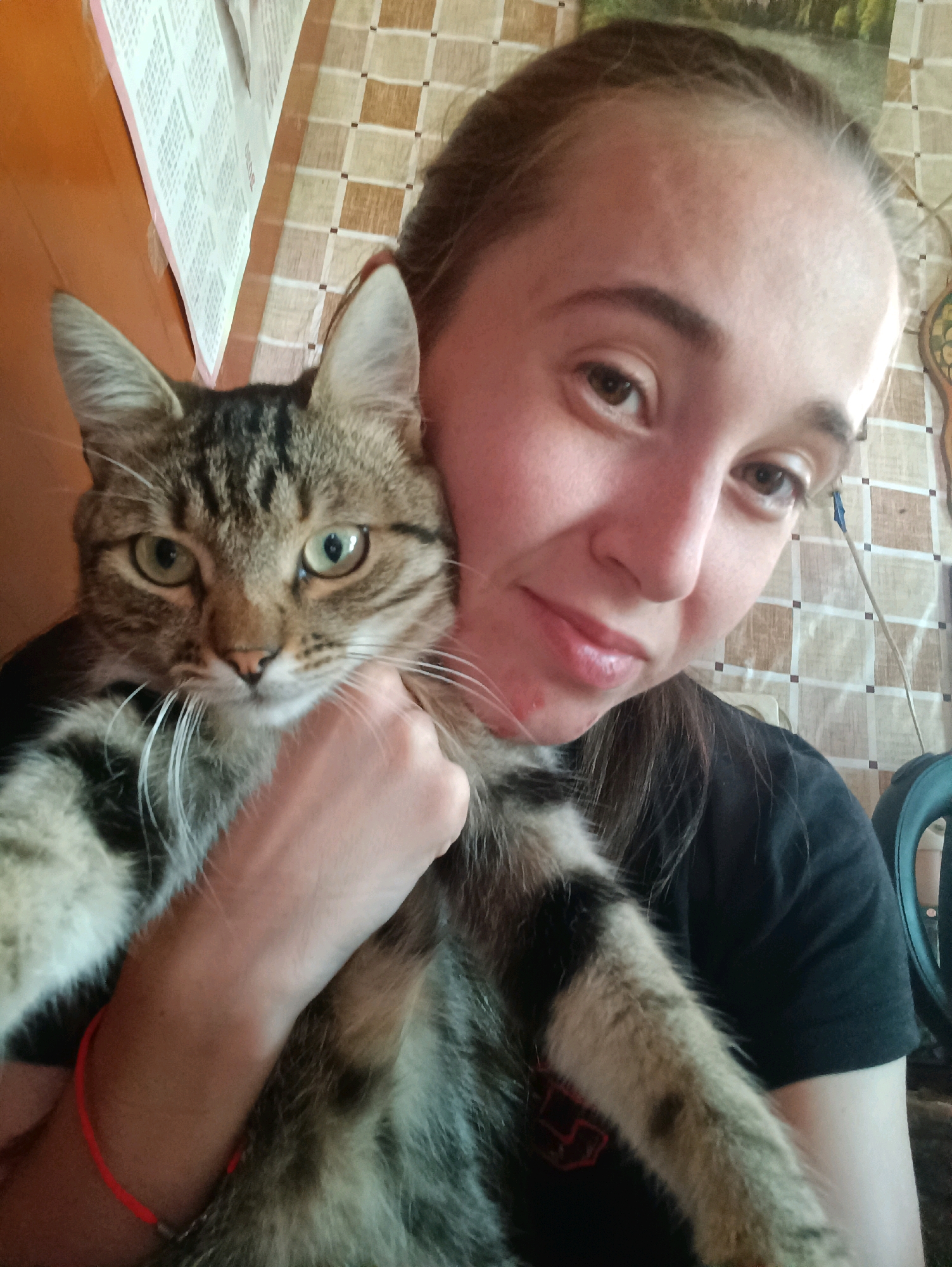 A young woman is taking a selfie with a tabby cat. The cat is held in the woman's left arm, with its head resting on her shoulder. The woman has brown hair pulled back, and is wearing a black t-shirt. The background appears to be indoors, with a wall covered in a checkered wallpaper and some papers hanging on the wall. The image is taken in Vypasne, Ukraine.
