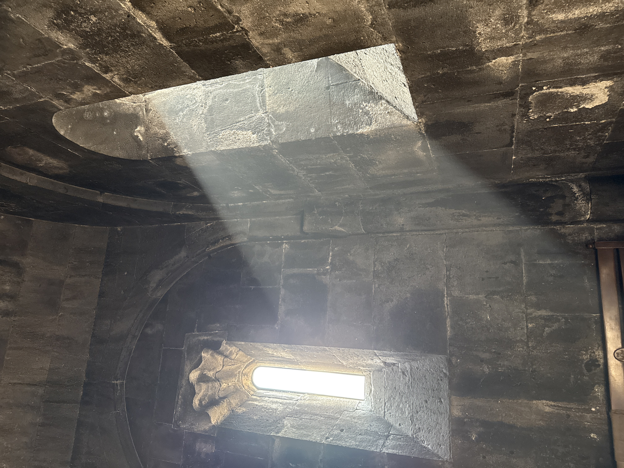 A sunbeam slices through the dusty interior of a stone building, illuminating the rough-hewn walls and a narrow, arched window. The light streams diagonally across the frame, creating a dramatic contrast between the brightly lit area and the shadowy corners. The stone blocks that make up the walls are large and irregular, showing signs of age and wear. The window, to the left, is tall and slender, with a decorative stone carving above it. The scene appears to be within the Geghard Monastery in Goght, Armenia, a UNESCO World Heritage site known for its rock-cut architecture.