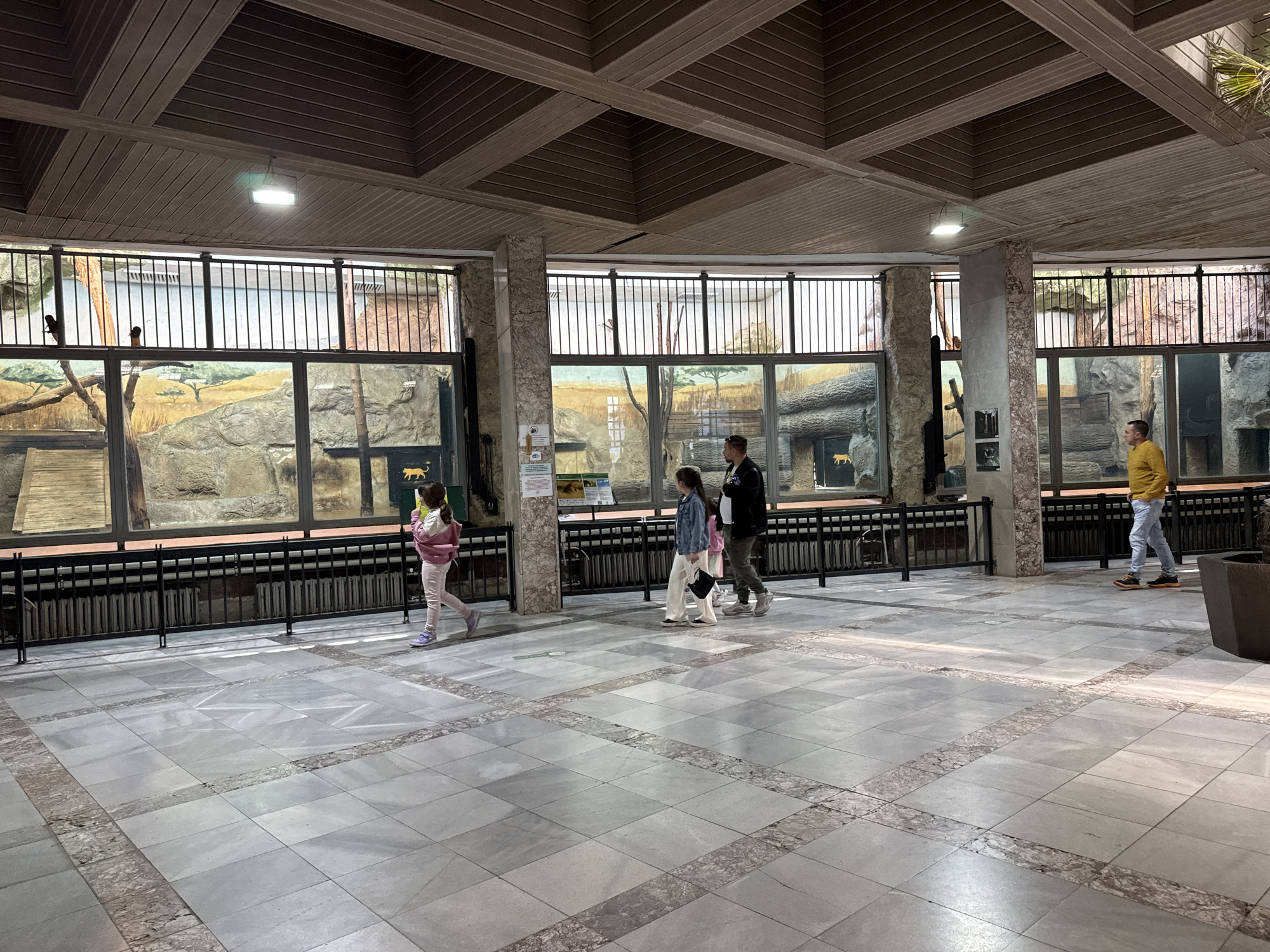 The image shows the interior of an exhibit hall, likely at the Sofia Zoo in Sofia, Bulgaria. The hall features a marble tile floor with inlaid stone patterns, and a coffered ceiling with recessed lighting. Large windows display habitat enclosures, possibly for big cats, with rocky terrain, painted backdrops, and animal silhouettes visible inside. Several people are walking through the hall, including a child in pink, a group of three people (two adults and a child), and an individual in a yellow sweater. Black metal railings run along the front of the windows, and potted plants are visible near the far end of the hall.