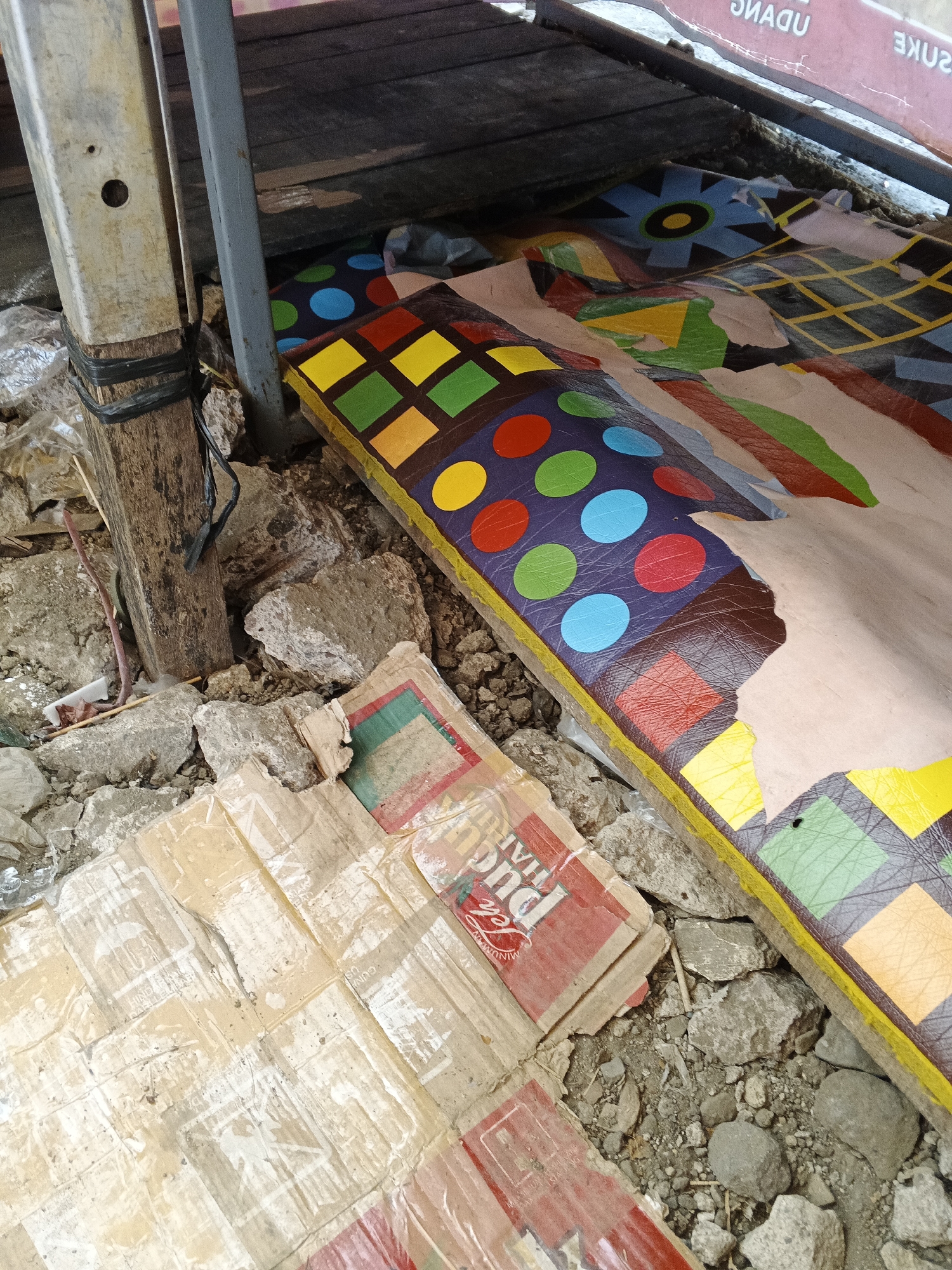 The image depicts a close-up view of a ground-level scene, likely an outdoor area in Sukabumi, Indonesia. A weathered wooden post, wrapped with black tape, stands on the left, adjacent to a pile of rubble and debris. A brightly colored, patterned surface, possibly a vinyl mat or tarp, is spread out, featuring geometric shapes like squares and circles in various hues of red, yellow, green, blue, and purple. This patterned surface is partially torn and appears to be lying on top of cardboard boxes. One visible box has red and green sections with some printed text. The ground is covered with small stones and dirt, consistent with a less-maintained or possibly a temporary outdoor setup. The overall impression is one of a makeshift or informal arrangement in a somewhat unkempt environment.