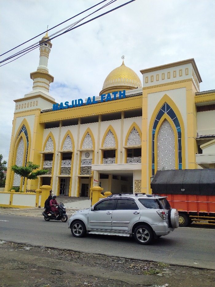 The image captures a vibrant street scene in Karanganyar, Indonesia, featuring the "MASJID AL FATH" mosque as its prominent backdrop. The mosque is a large, brightly colored building with a facade primarily in yellow and white, accented with blue details on its arched window frames and the mosque's name. It features a tall minaret on the left with a golden finial, and a large golden dome at its center, also topped with an Islamic crescent and star. Intricate white patterns fill the arched windows and panels along the mosque's lower level.
In the foreground, a two-lane asphalt road runs horizontally. On the left side of the road, a motorcycle with two riders, one wearing a red top, is visible, appearing to move past the mosque. To the right of the motorcycle, a silver SUV, possibly a Toyota Rush given its distinctive spare tire mounted on the rear door, is also on the road, heading in the same direction. Further to the right and slightly behind the SUV, a large red truck with a black tarp covering its cargo area is also seen on the road.
The sky is overcast, suggesting a cloudy day, though the lighting is bright. Overhead, several power lines stretch across the frame, partially obscuring the sky above the mosque. The immediate roadside in the foreground shows some gravel and uneven terrain. The overall impression is a bustling daytime scene in an urban or semi-urban setting, with a striking religious building as a central focal point.
