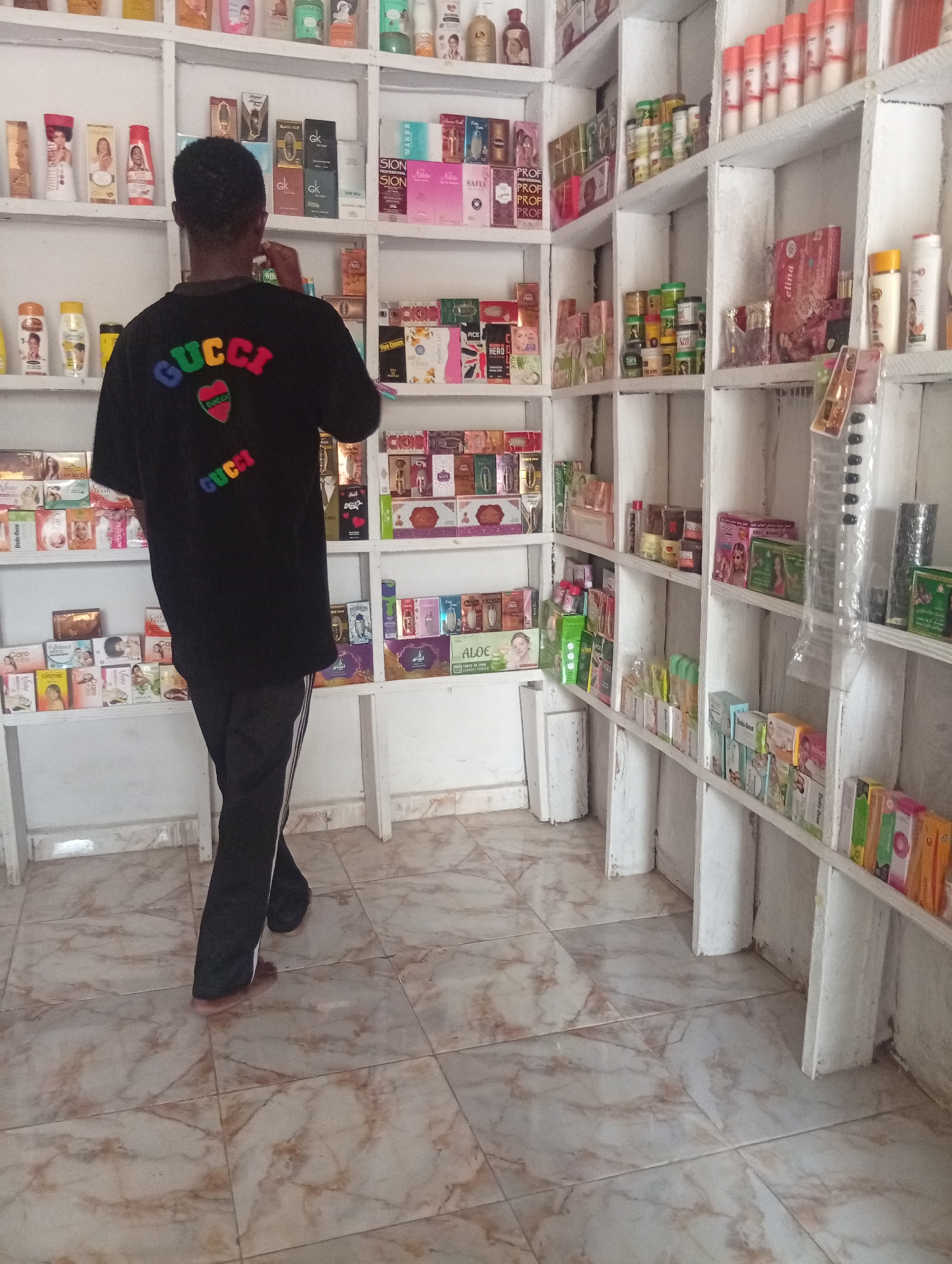 An indoor scene depicts a retail store, likely a beauty supply or personal care shop, located in Unknown, Unknown. A single individual, seen from behind, stands centrally in the frame, facing shelves laden with various products. The person wears a black short-sleeved t-shirt featuring a multi-colored "GUCCI" logo and a striped heart graphic on the back, along with black athletic pants with white side stripes. The individual is barefoot, with the soles of their feet visible on the tiled floor. Their right hand is raised to their mouth or chin area.
The store is furnished with multiple tiers of white, unfinished wooden shelves lining the walls. These shelves display a wide array of products, predominantly cosmetics, skincare items, perfumes, and lotions, in various bottles, jars, and cardboard boxes. Visible brands include "WHITE SECRET," "GK" (G.K. Paris), "SION," "Nilam," "SAFIA," "PROFI," "ELINA," "ZARA" (perfumes), "DREAM," "Vaseline," "Fair & Lovely," "Sky," "Veet," "Folebac," "Caro White," "Charitone," "Vivia," "ALOE," "ALIYA," "NAOMI," "Oud," "HERO D," and "Dudu-Osun." Many product boxes feature images of people, often women. On the right side, a section of the top shelf holds numerous uniform orange and white cylindrical containers. Below this, a transparent plastic package containing small black items hangs from a shelf. The floor is covered with light-colored, marbling-patterned ceramic tiles in shades of beige and brown. The walls behind the shelves are painted white.